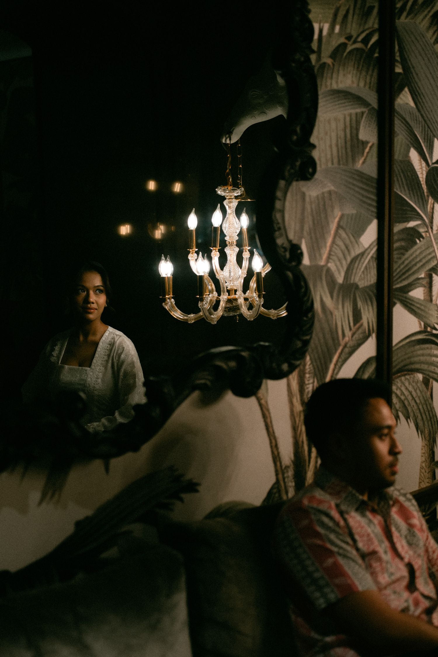 Moody interior scene with ornate crystal chandelier and decorative mirror casting warm ambient lighting in dark room.