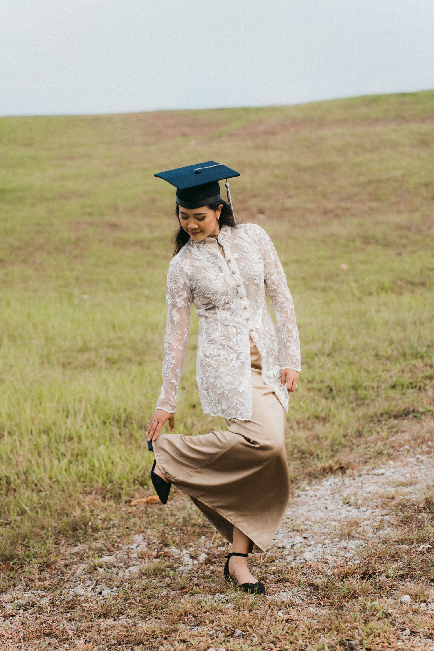 Graduate in white lace dress and cap sits thoughtfully in a grassy field during graduation photoshoot.