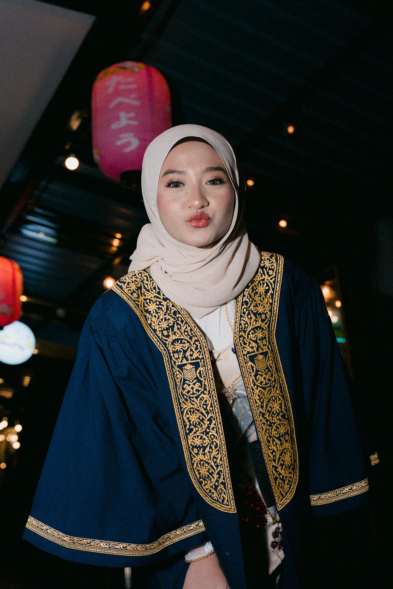 Young woman in hijab and embroidered navy robe posing indoors with bokeh lights and red lantern background.
