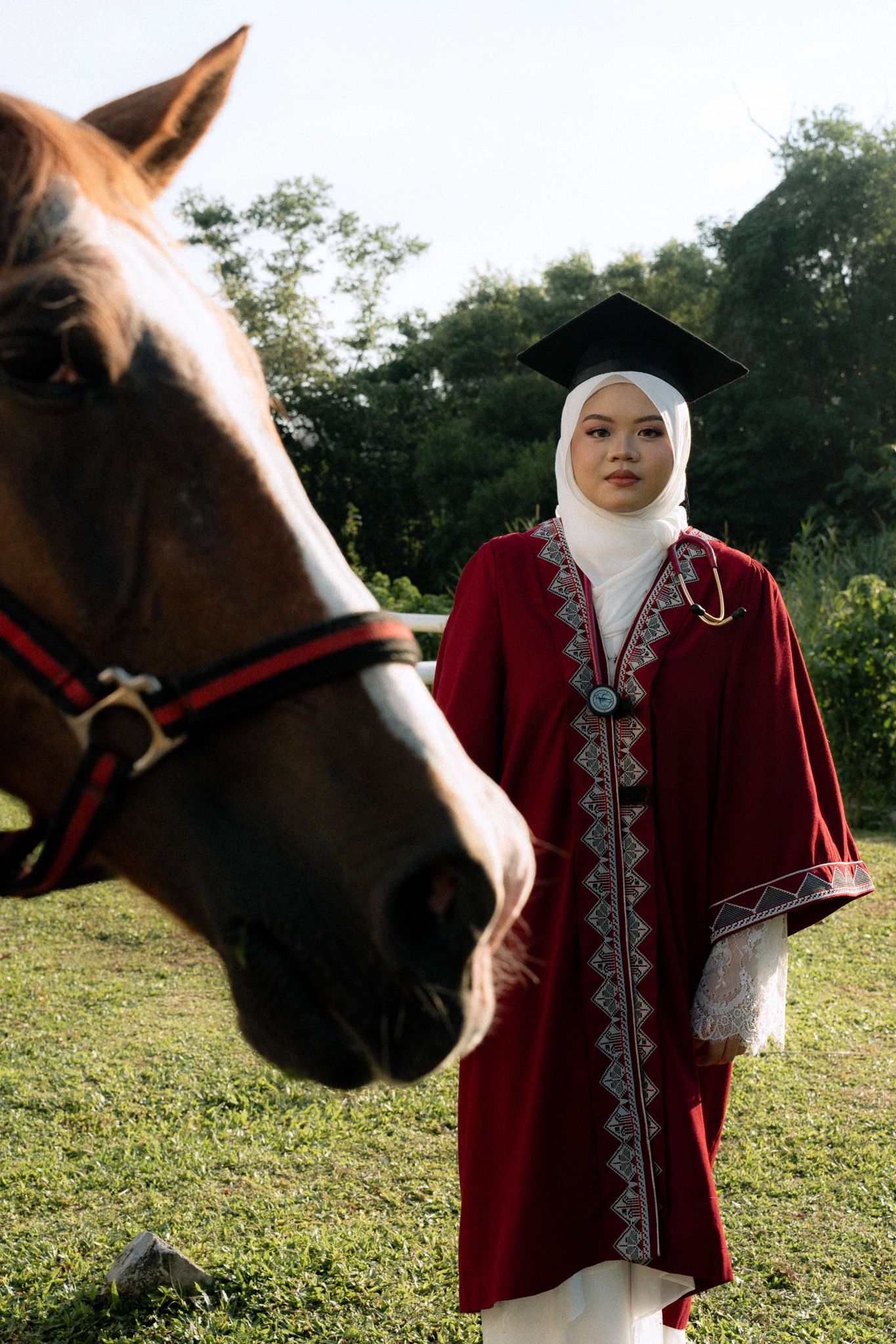 Muslim woman in red graduation gown and hijab holds diploma beside a brown horse outdoors.