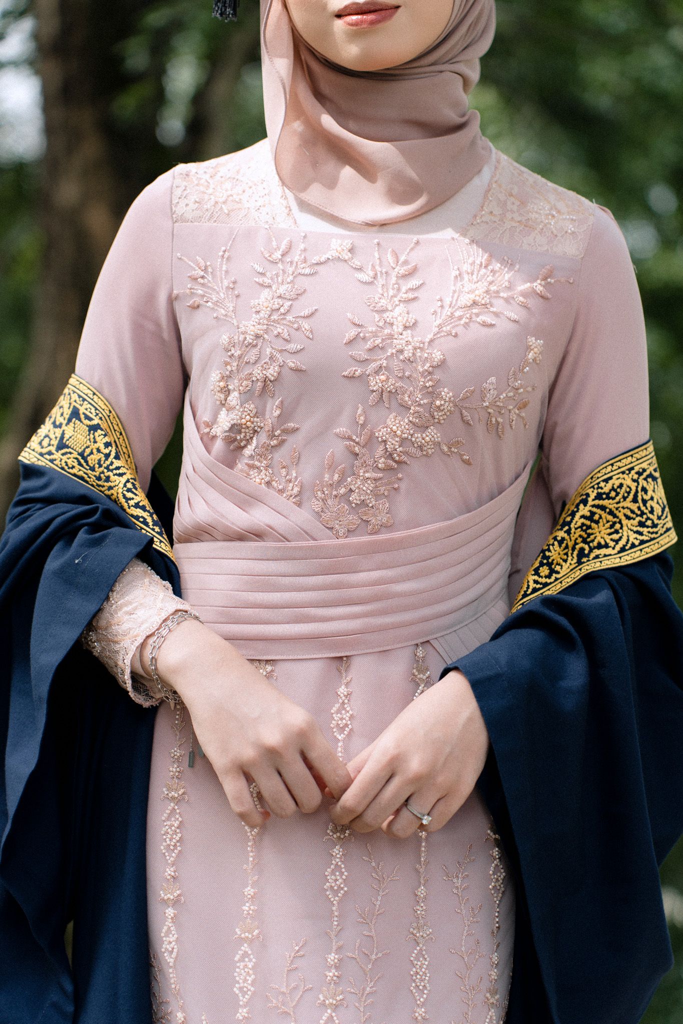 Woman wearing an embroidered pink modest dress with floral details and a hijab, paired with a navy blue cardigan with gold trim.