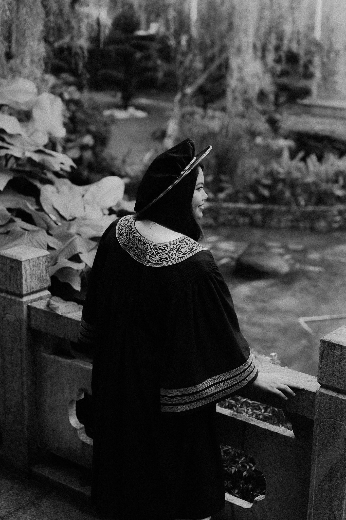 Black and white photo of a smiling woman in traditional embroidered dark attire standing by ornate stone railing in a garden.