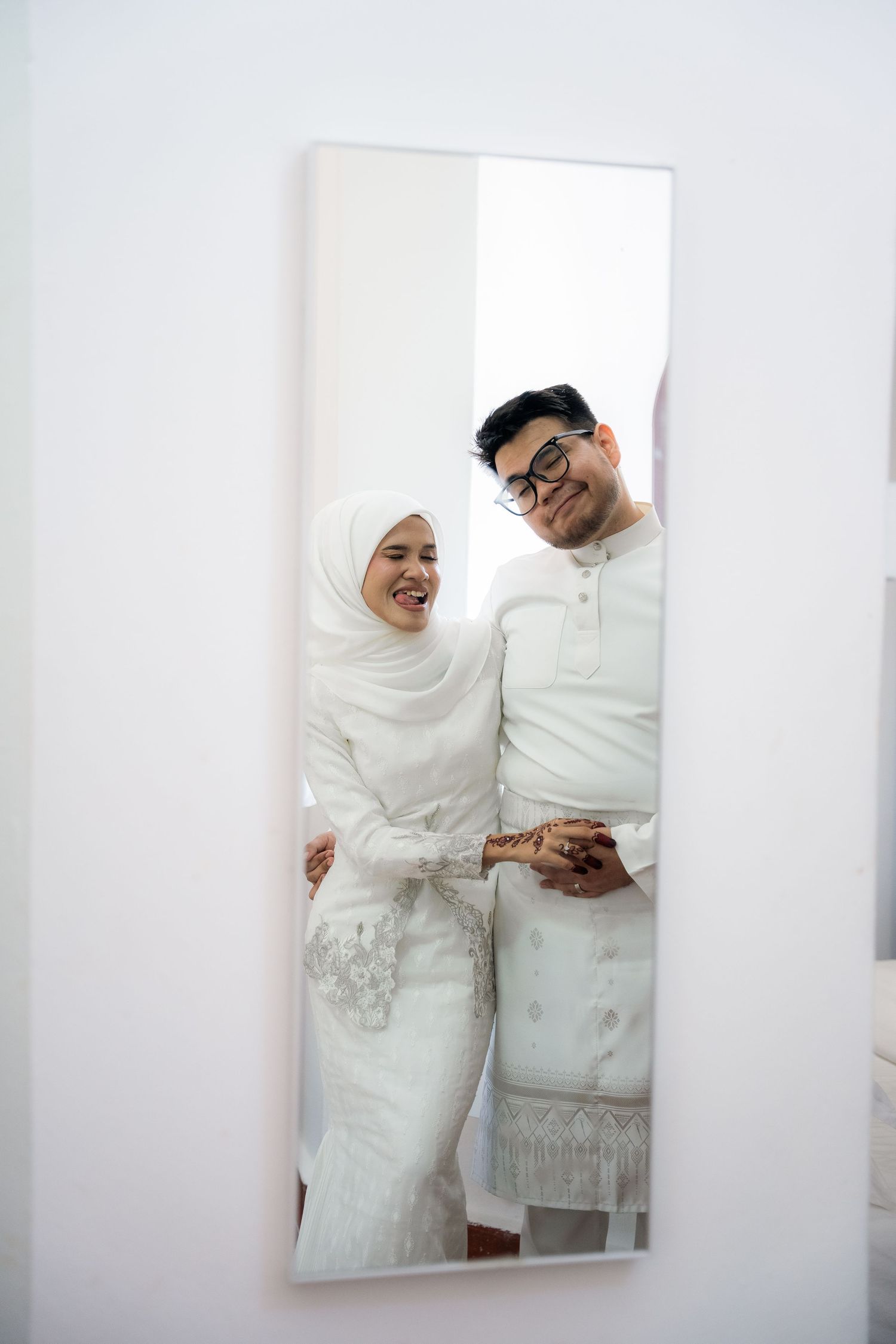 A couple shares joyful laughter together while wearing traditional white wedding attire against a bright white backdrop.
