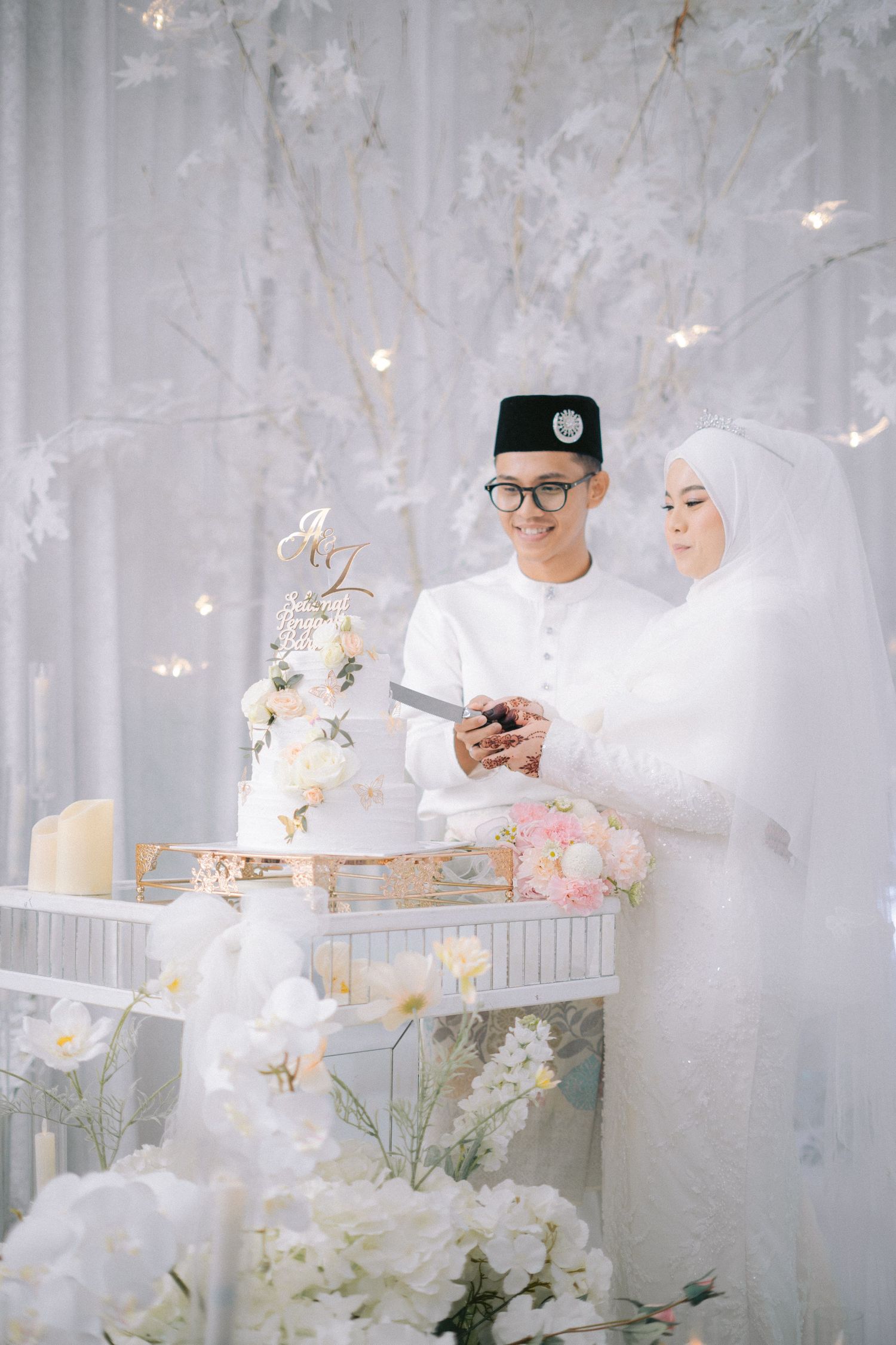 Traditional Malay couple in white attire stand by elegant wedding cake display with floral decorations and string lights.