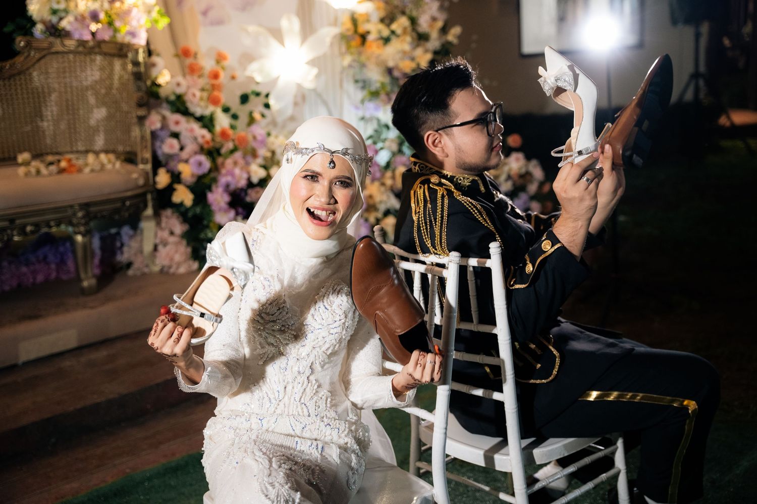 A couple in rocker chairs laughs joyfully at a vintage-themed wedding celebration with floral decorations in the background.
