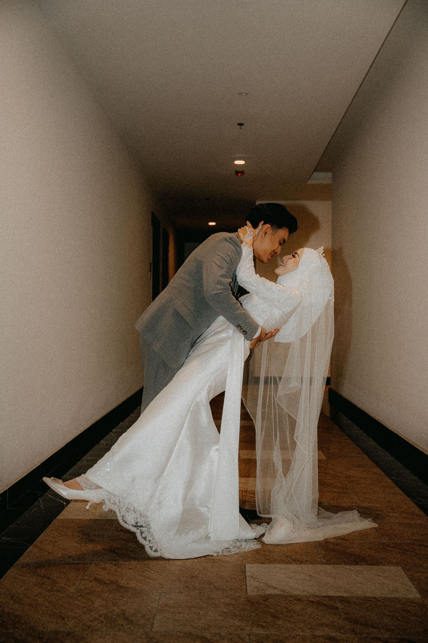 A romantic wedding couple shares a dip kiss in a hotel hallway, with the bride's flowing veil and white gown trailing behind.