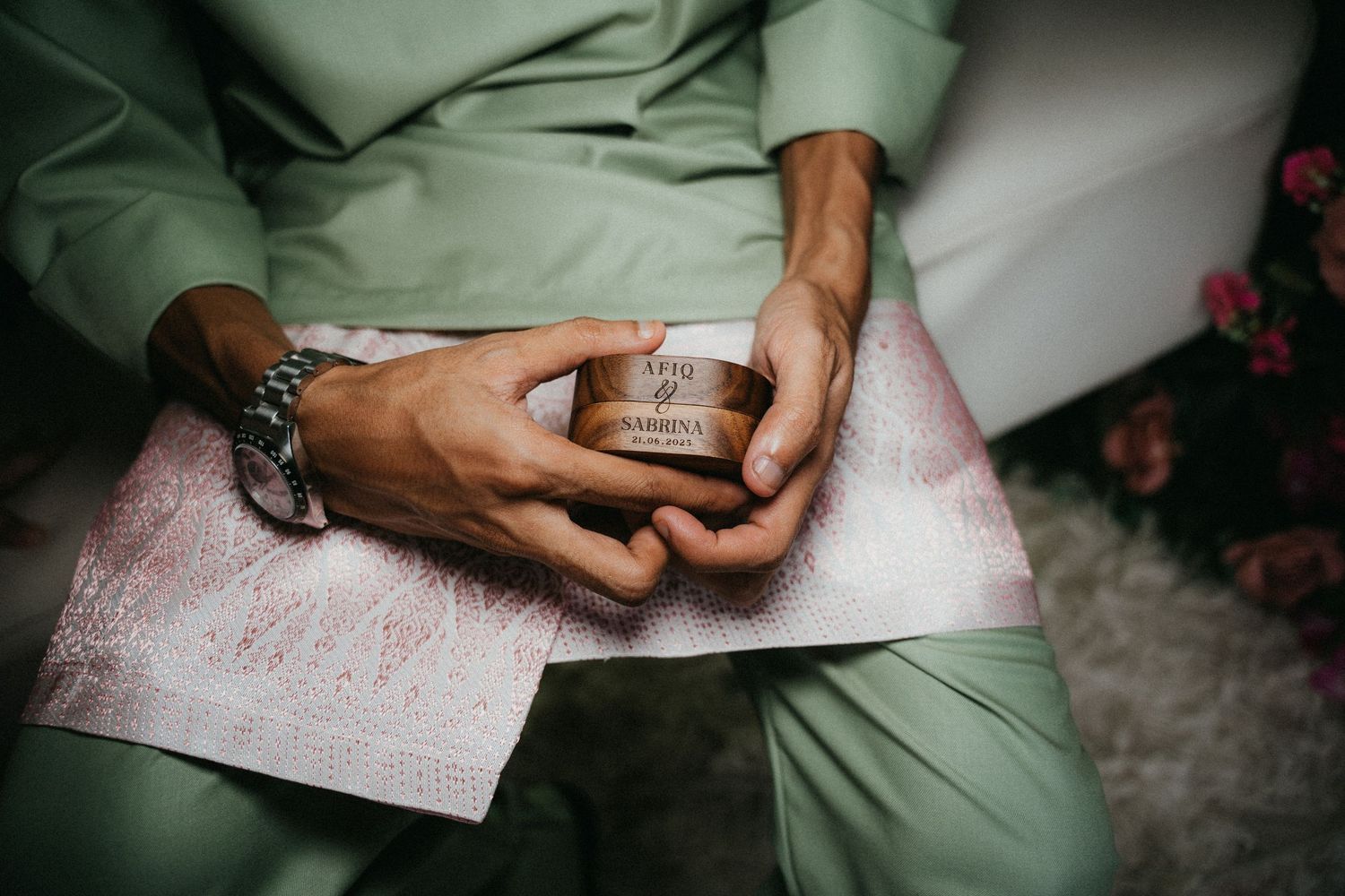Hands holding an engagement ring box while seated, wearing a mint green outfit against a light background.