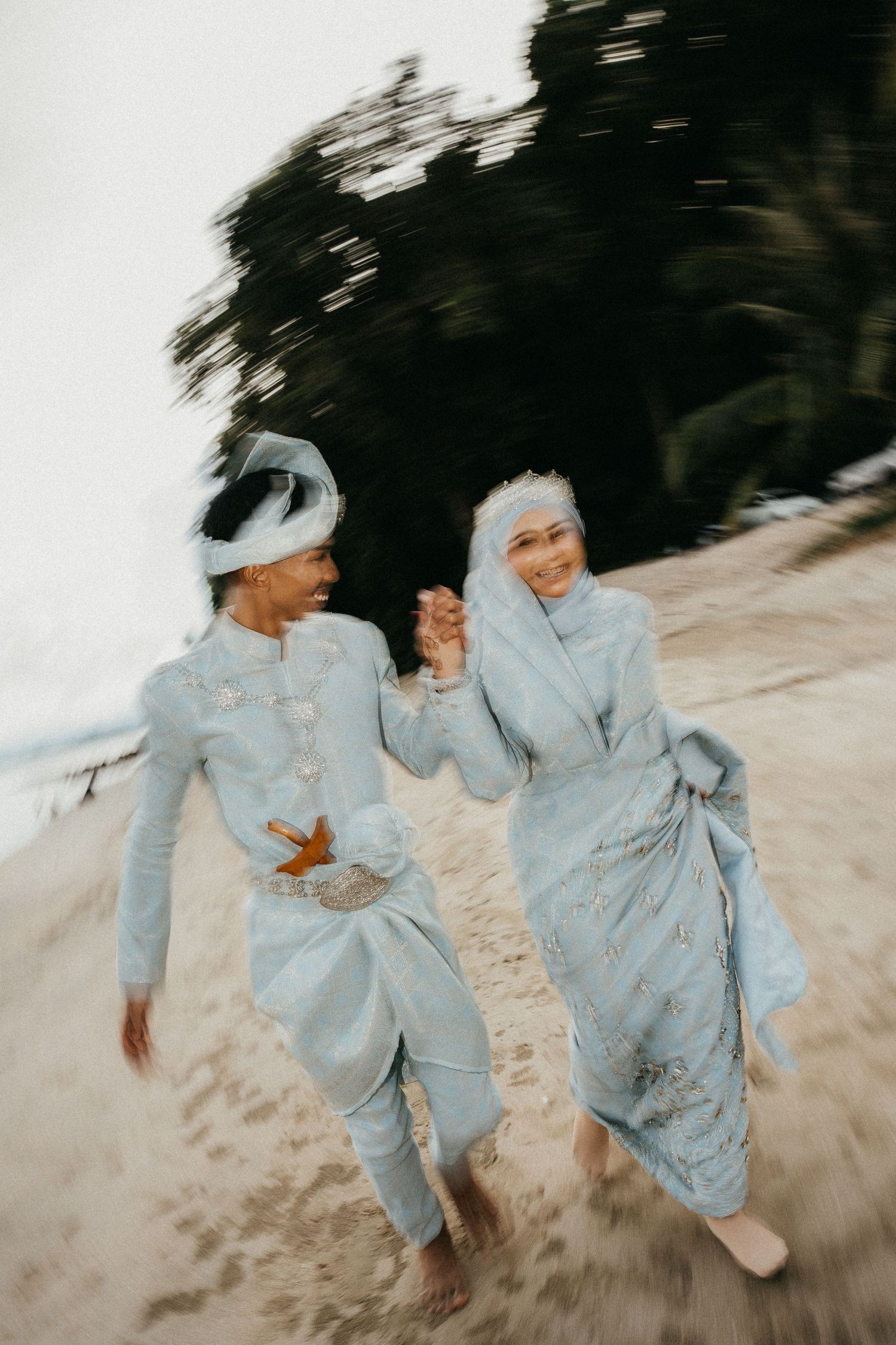 Two people in light blue traditional attire run joyfully along a sandy beach while holding hands.