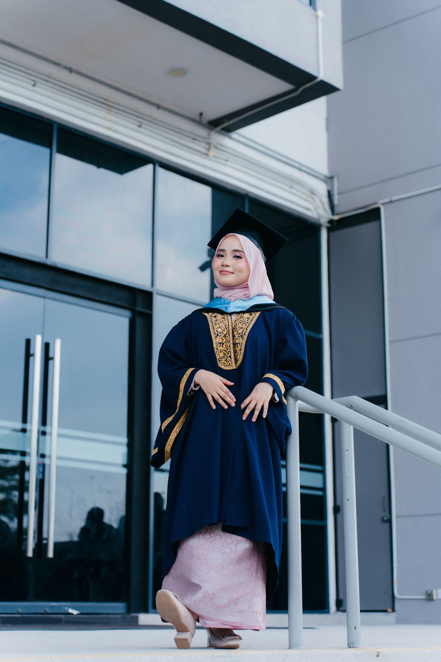A person in graduation attire sits on white steps outside a modern building with glass windows.
