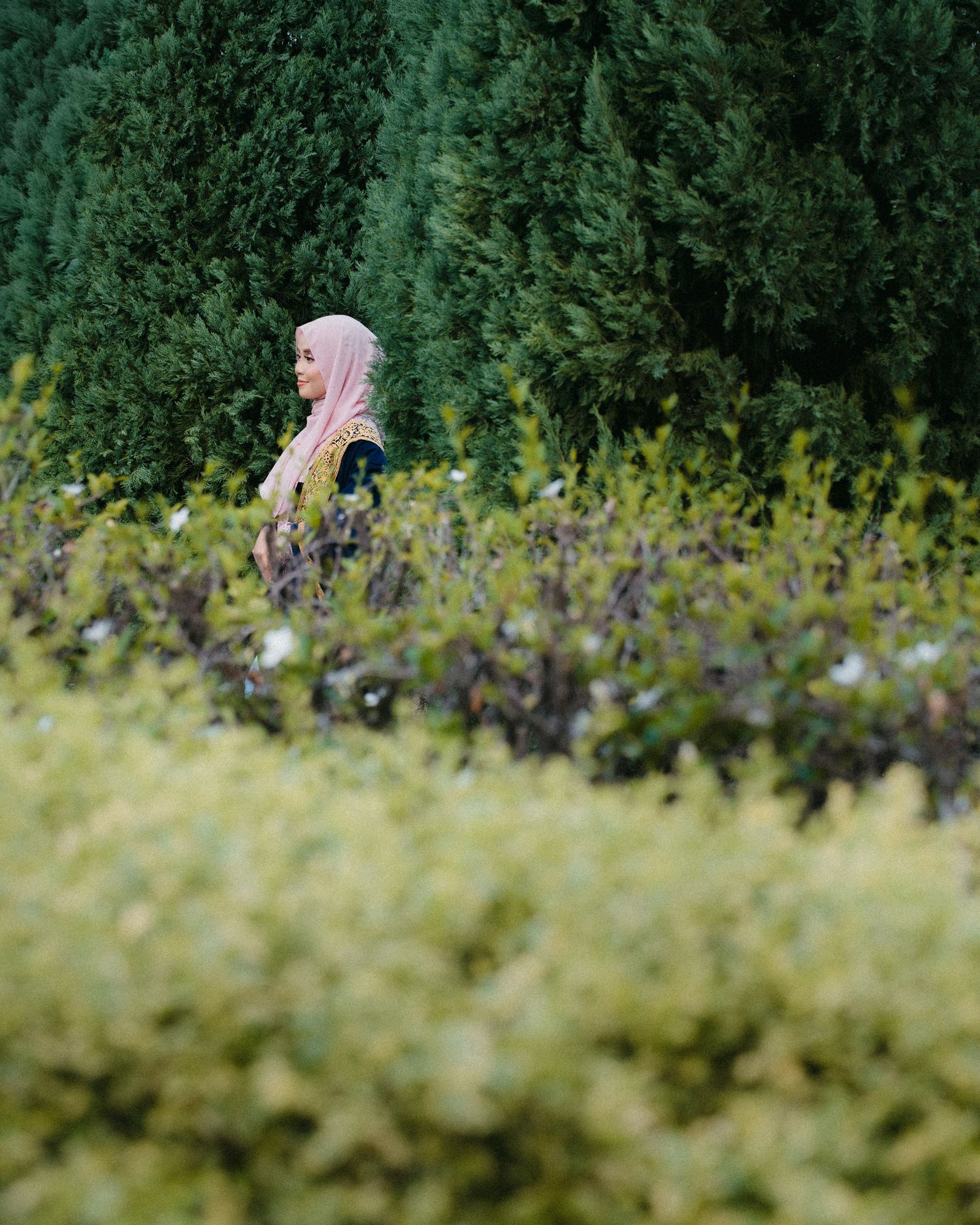 A person in a pink hijab is seen through blurred foliage in a garden setting.