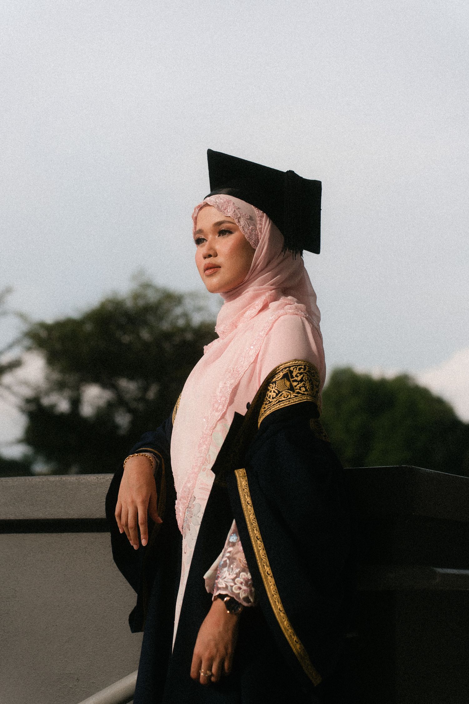 Graduate wearing black cap and gown with pink hijab poses for graduation photos outdoors at sunset.