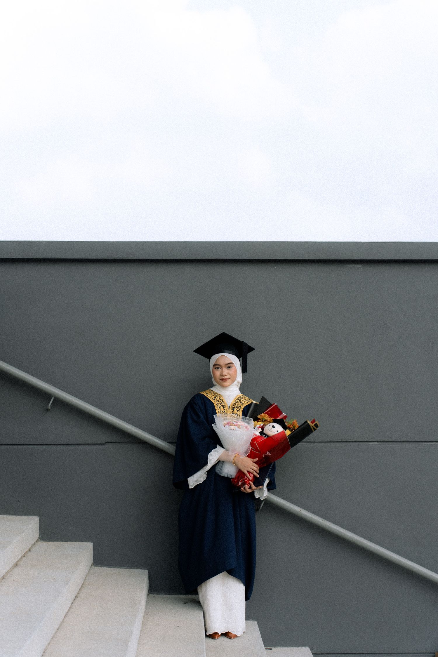Graduate in cap and gown holds a diploma and teddy bear while posing against a minimalist gray wall.