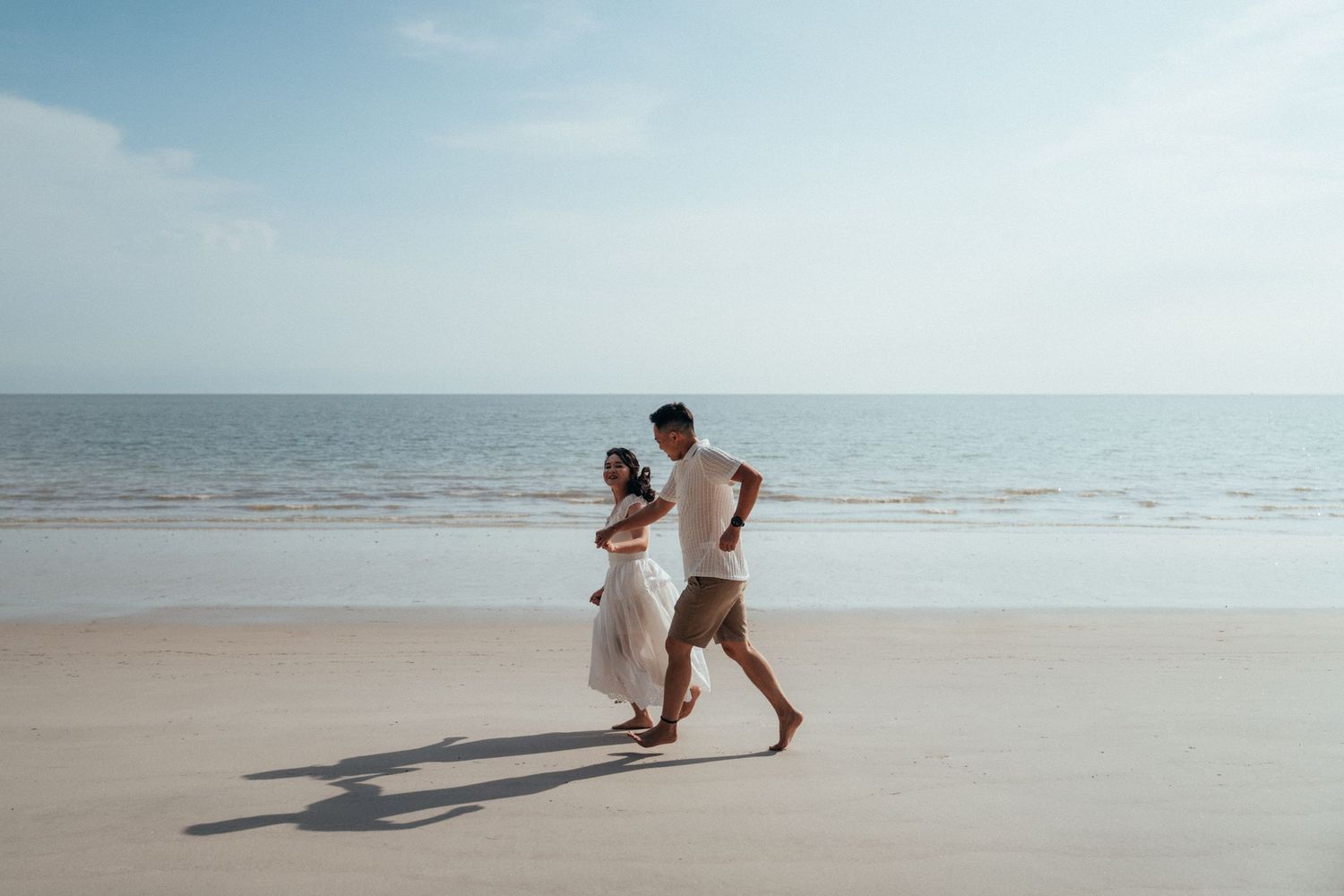 A couple runs joyfully together on a sandy beach with ocean waves in the background.
