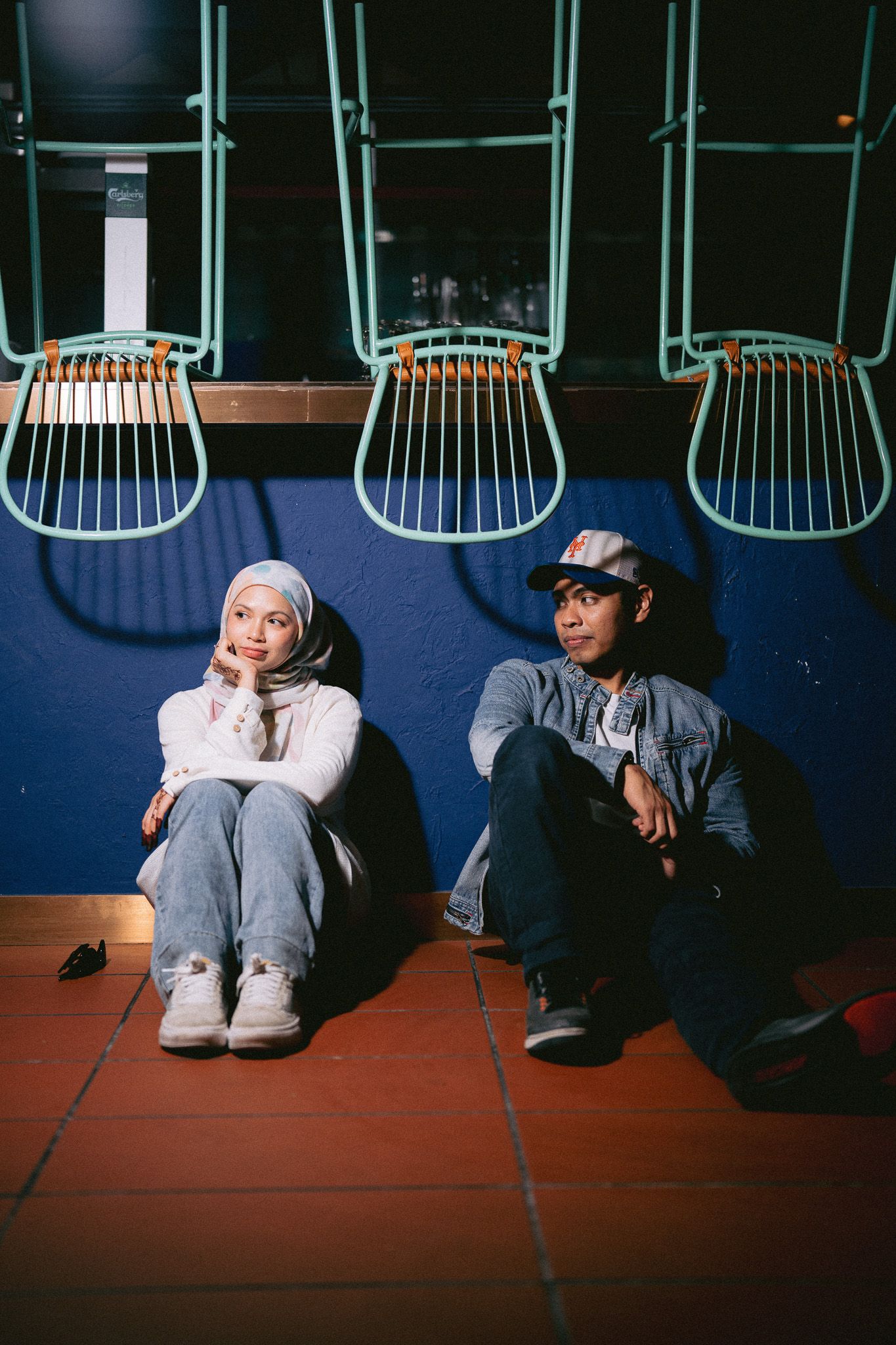 Two people sitting on a tiled floor against a blue wall with suspended chairs, one wearing a hijab and the other a cap.