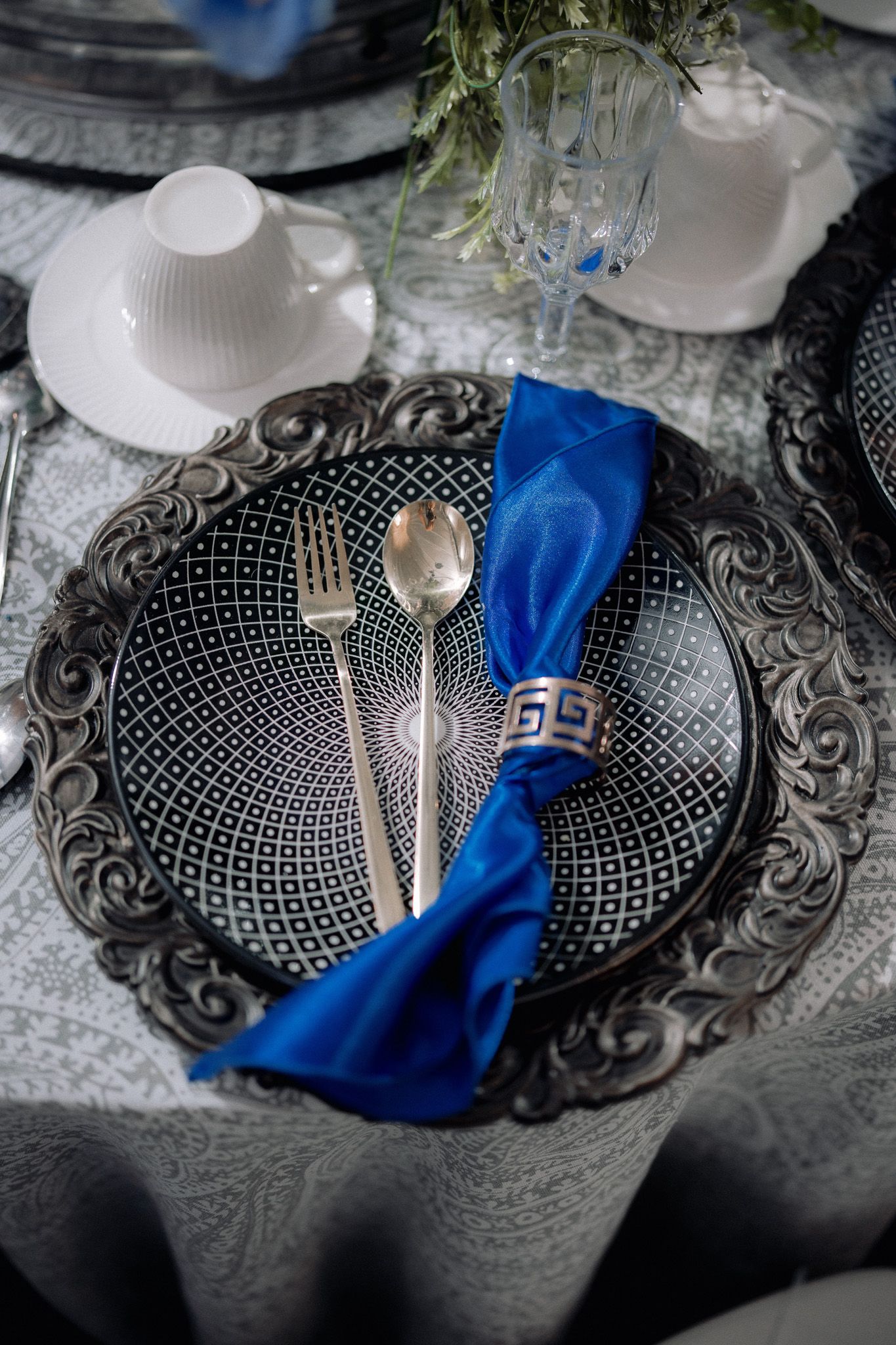 Elegant table setting with ornate silver charger plate, blue napkin, silverware, and patterned dinnerware on white tablecloth.