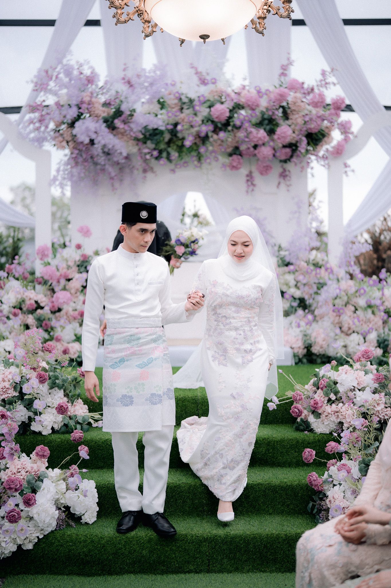 Malay couple in traditional white wedding attire walking through floral arch with pink and white flowers.