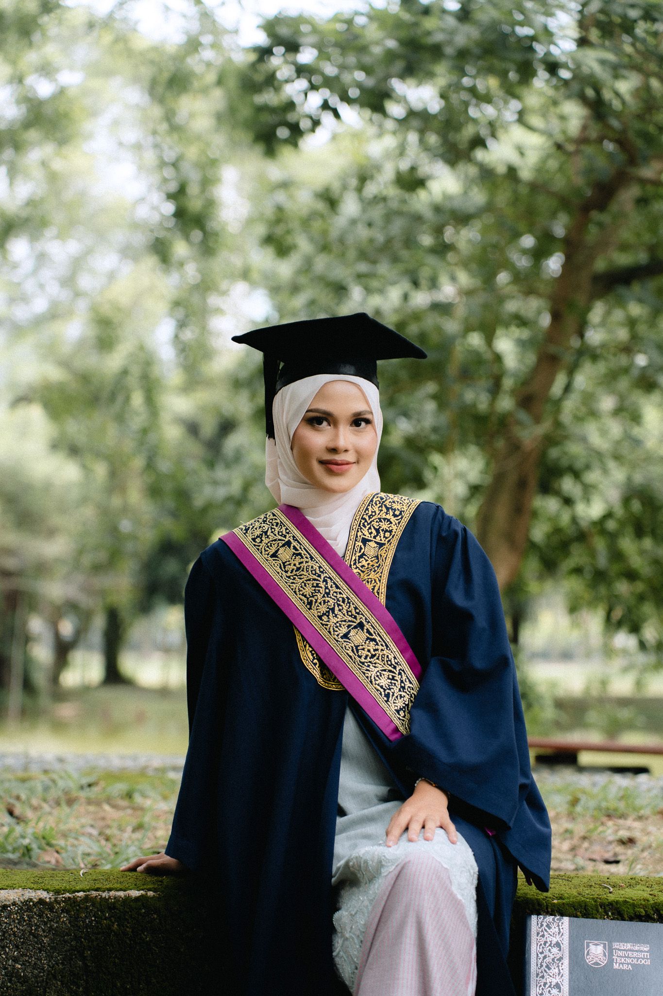 Muslim woman in graduation gown and hijab smiling outdoors in a park setting.