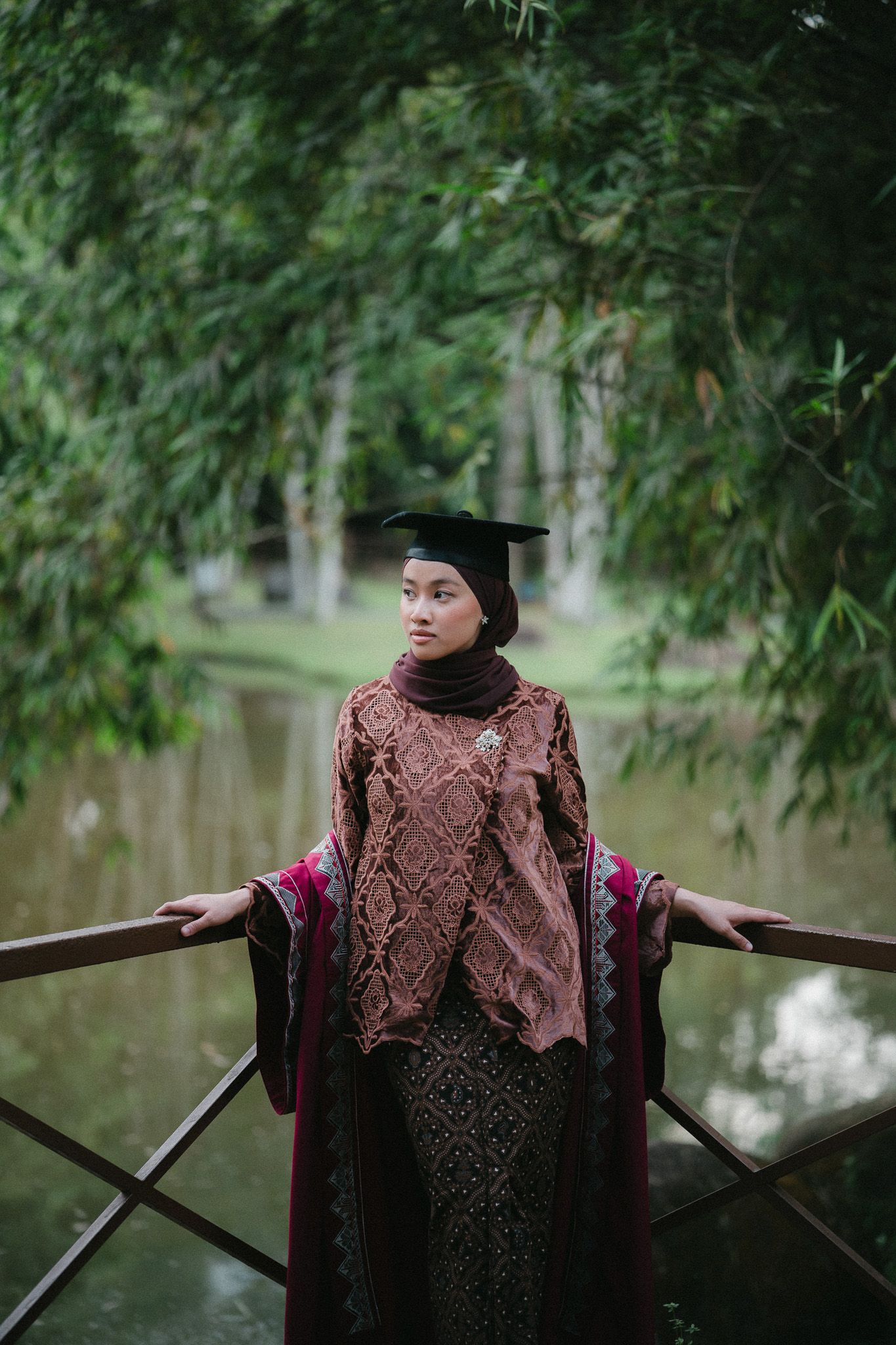 A young woman in a hijab and graduation cap poses on a wooden bridge surrounded by lush green trees.
