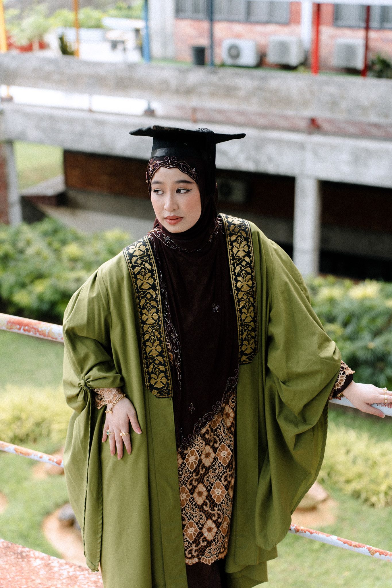 Muslim woman in green graduation robe and mortarboard cap with hijab posing outdoors on campus grounds.