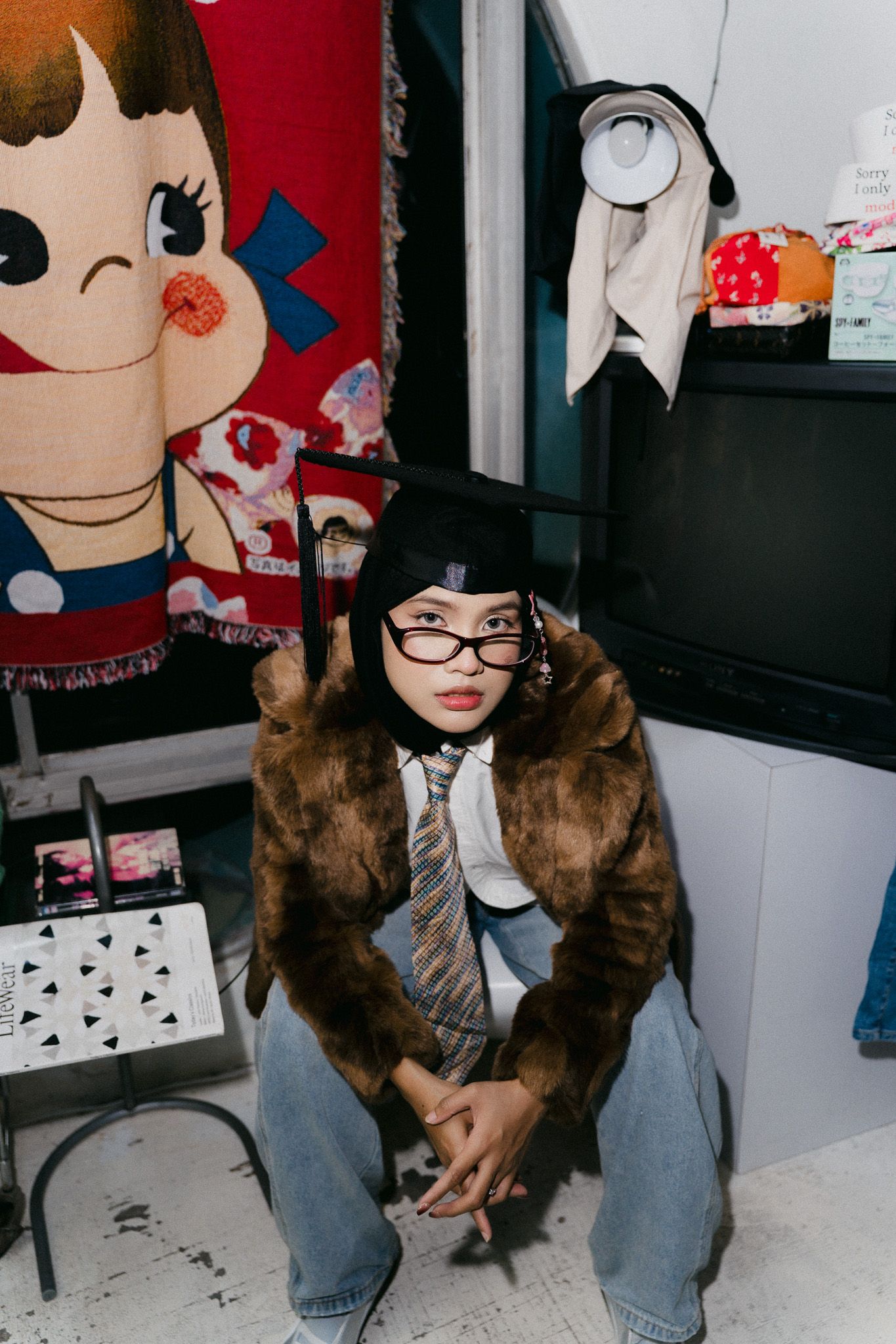 A young person in a fur coat, tie, glasses, and black beret crouches in a room decorated with colorful anime-style tapestries.