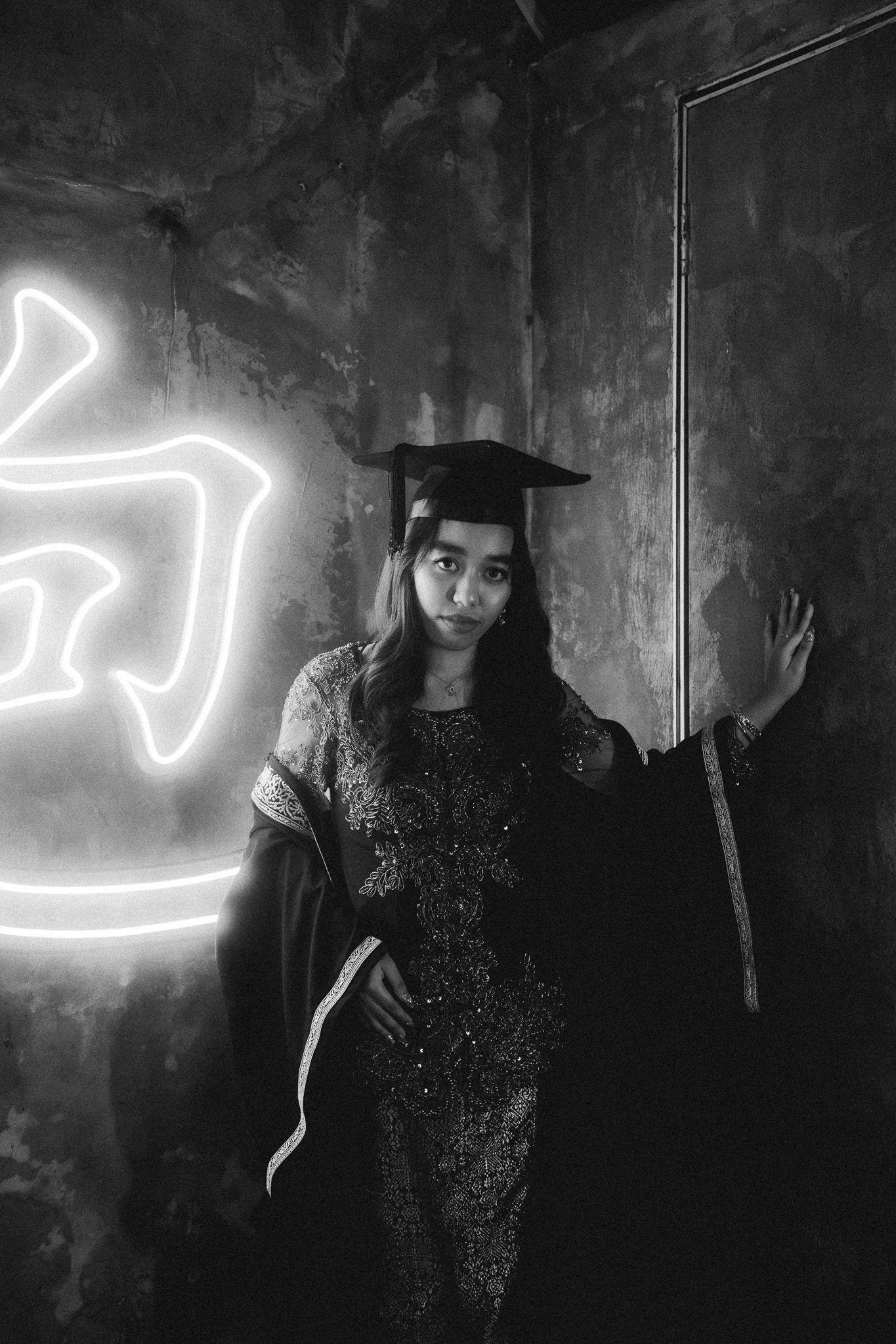 Black and white photo of a young woman in graduation cap and gown posing near a glowing neon sign.