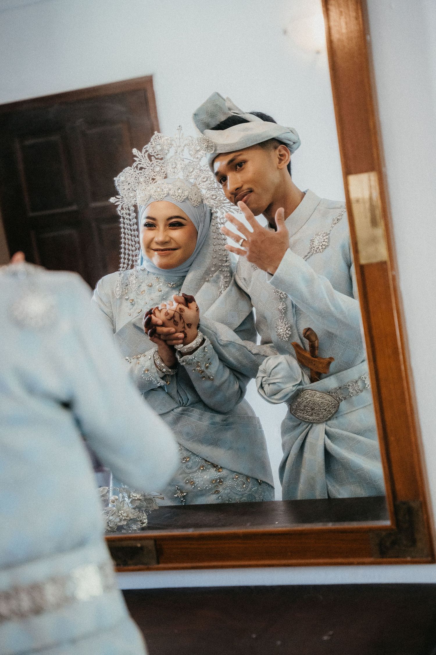Reflection in mirror shows a couple in traditional Malay wedding attire with silver and grey formal clothing and headpieces.