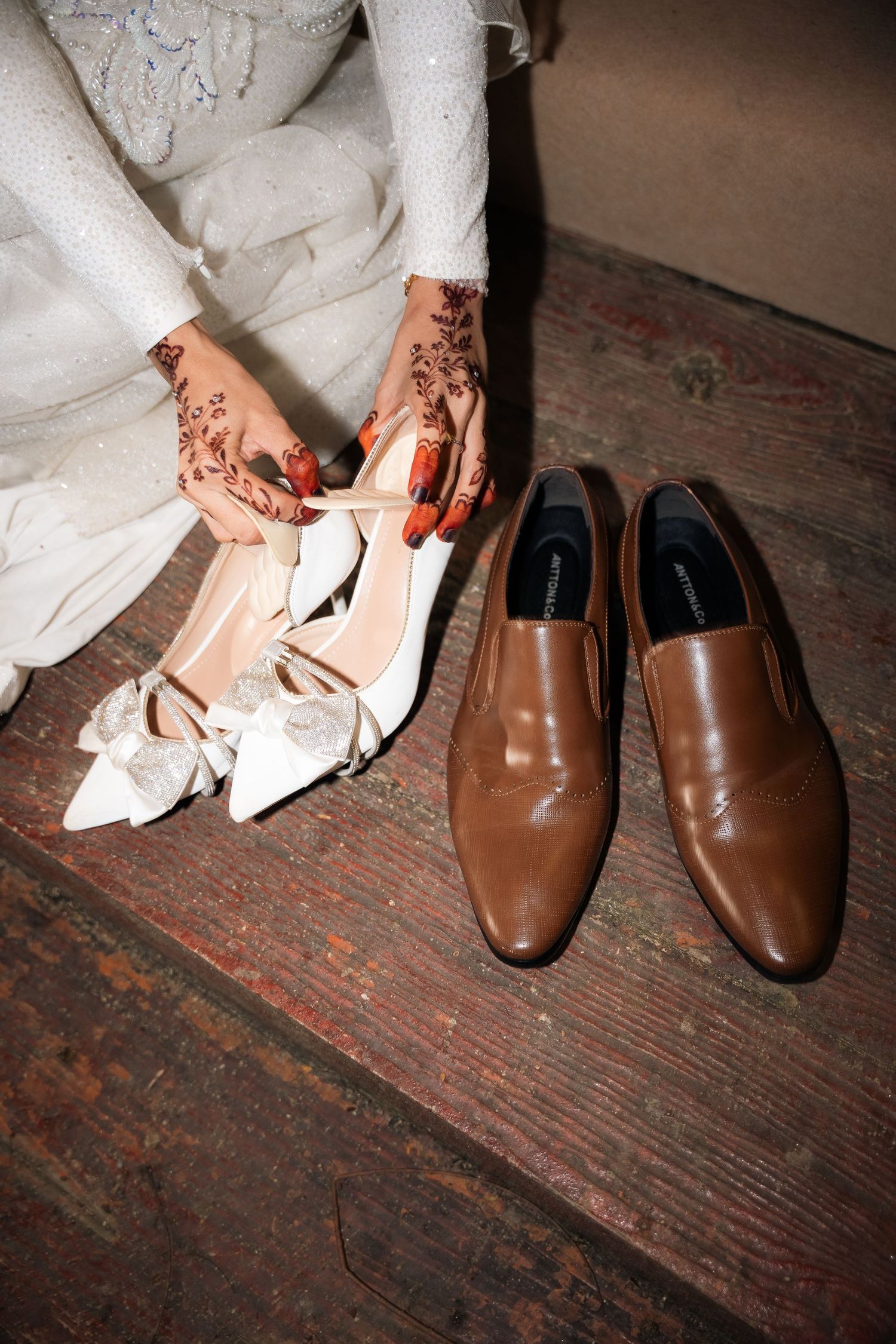 Wedding shoes side by side on wooden floor showing brown leather groom's loafers and white bridal heels with henna decorated hands.