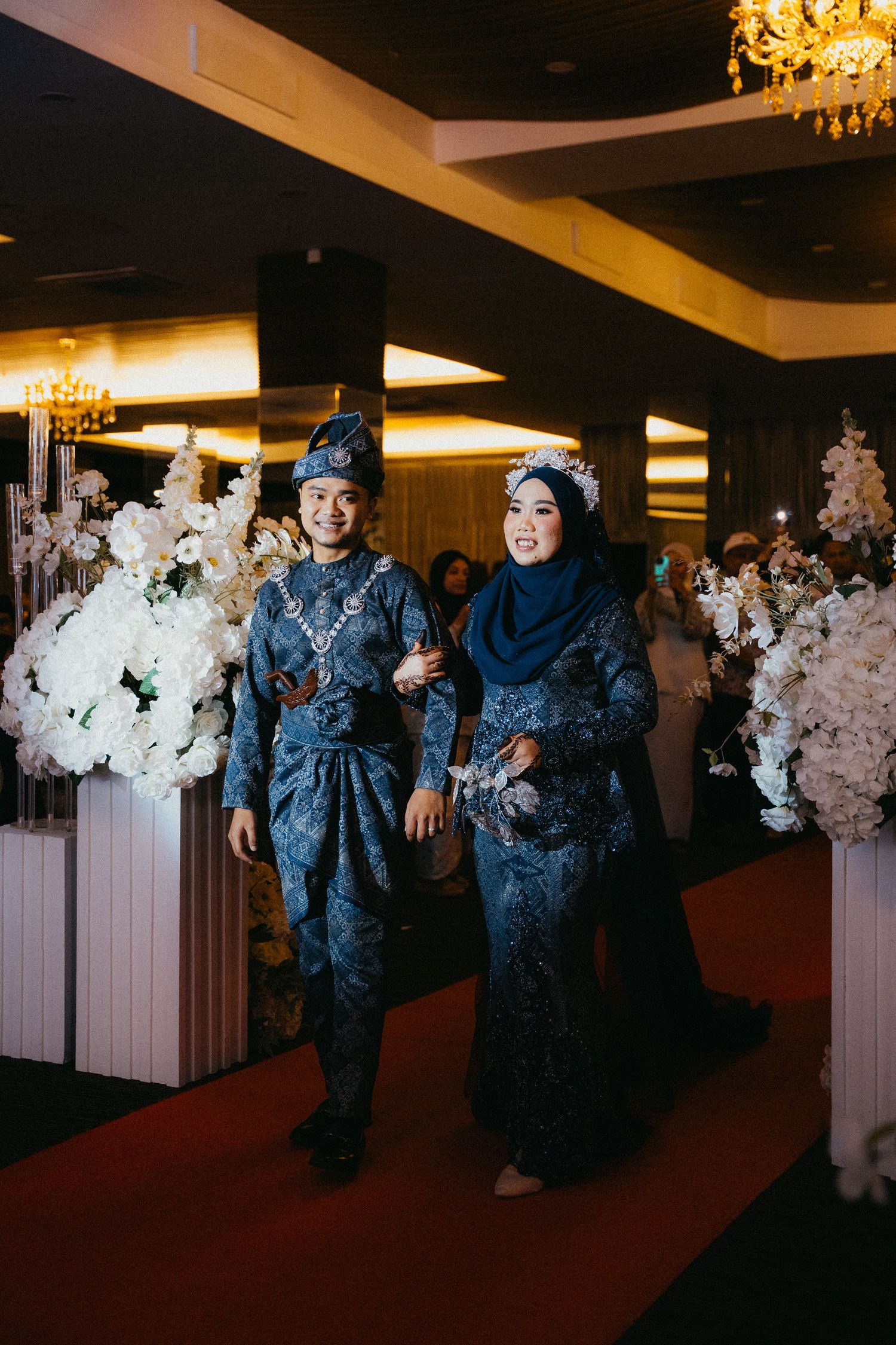 Ring bearers in matching navy blue suits walk down the aisle at an elegant wedding reception with white floral decor.