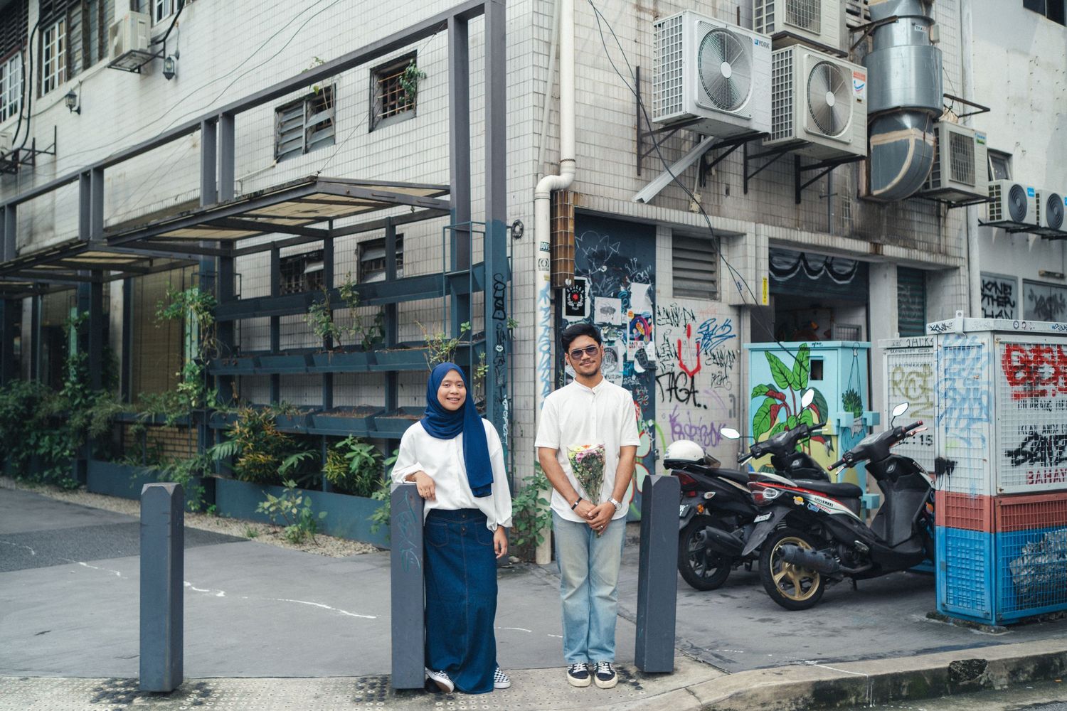 Two people stand near parked motorcycles on an urban street with air conditioning units.