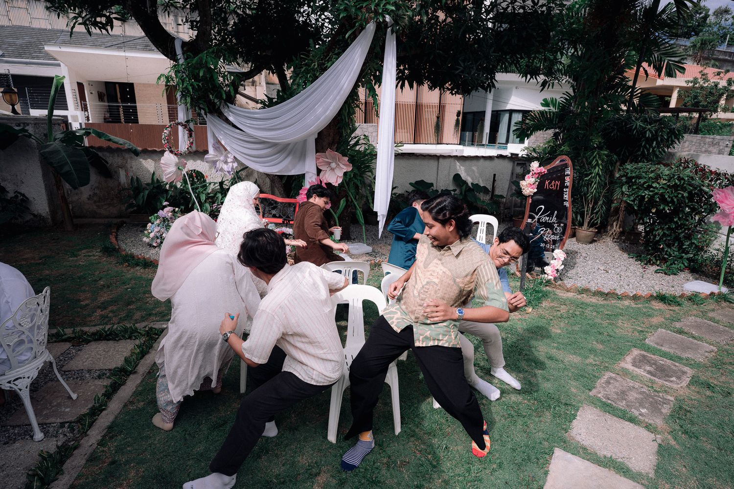Group of people gathered around a white table in a lush backyard garden with decorative draping overhead.