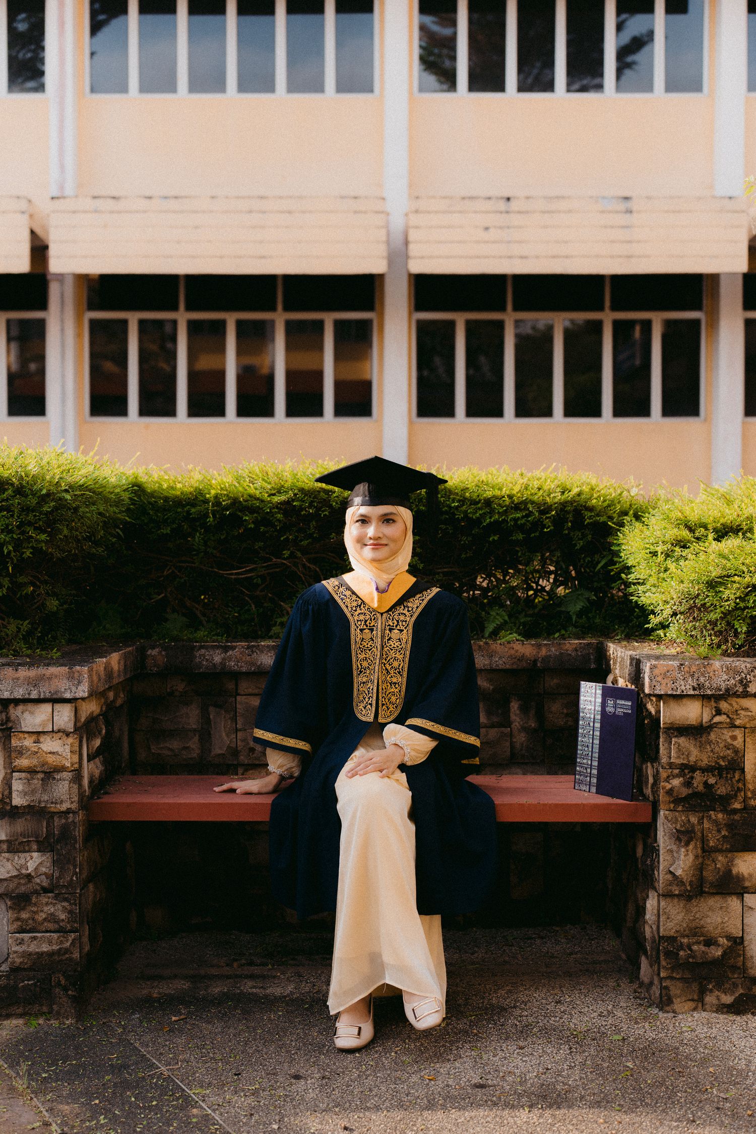 A graduate wearing traditional regalia and mortarboard sits on a stone bench near landscaped shrubs outside a building.