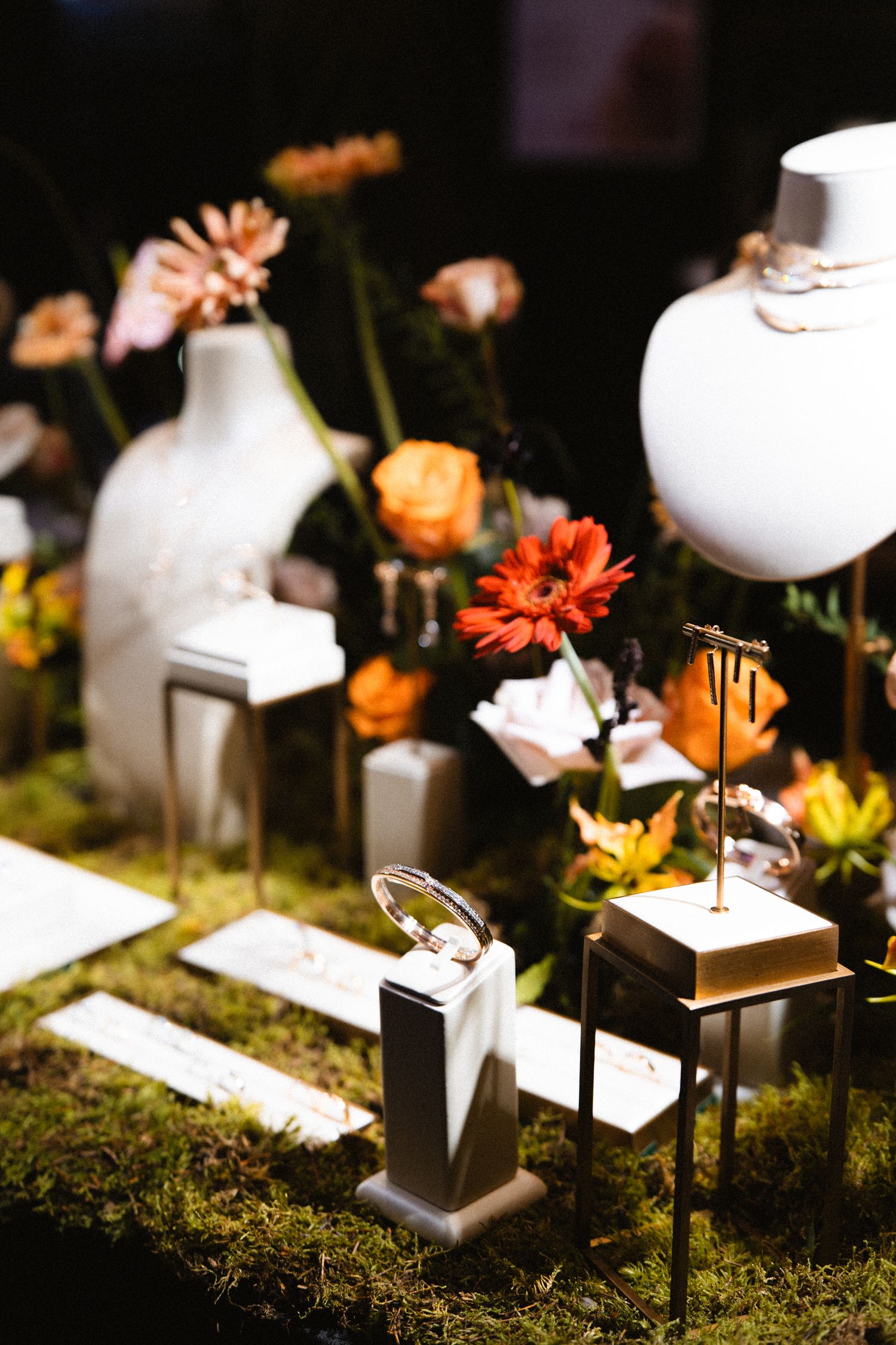 Jewelry display with orange and pink flowers on moss-covered pedestals in dramatic lighting.
