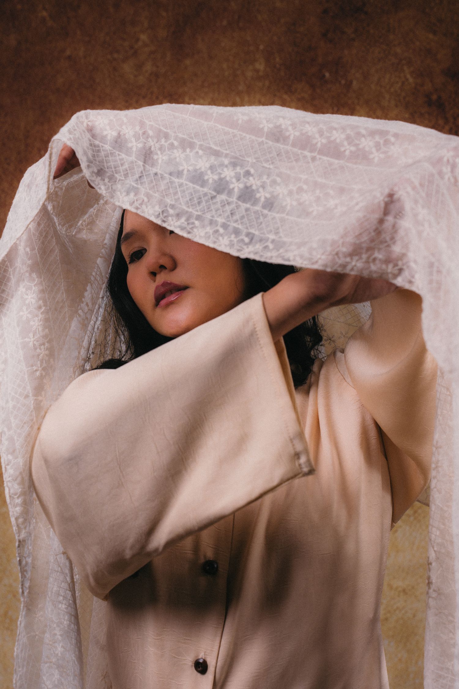 A person in beige clothing poses artistically with sheer lace fabric against a textured brown backdrop.