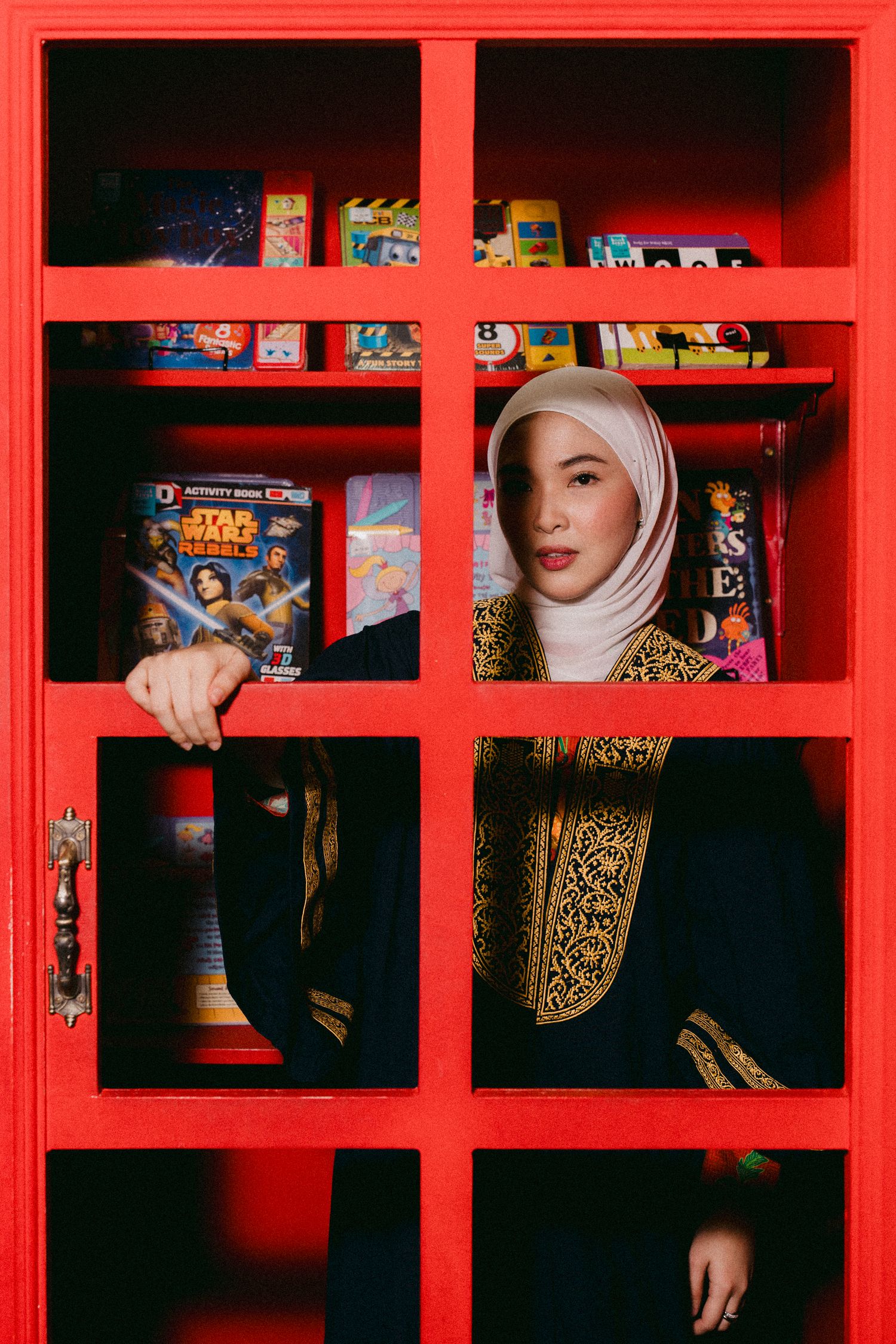 Graduate in hijab and embroidered robe poses in red framed window with books visible.