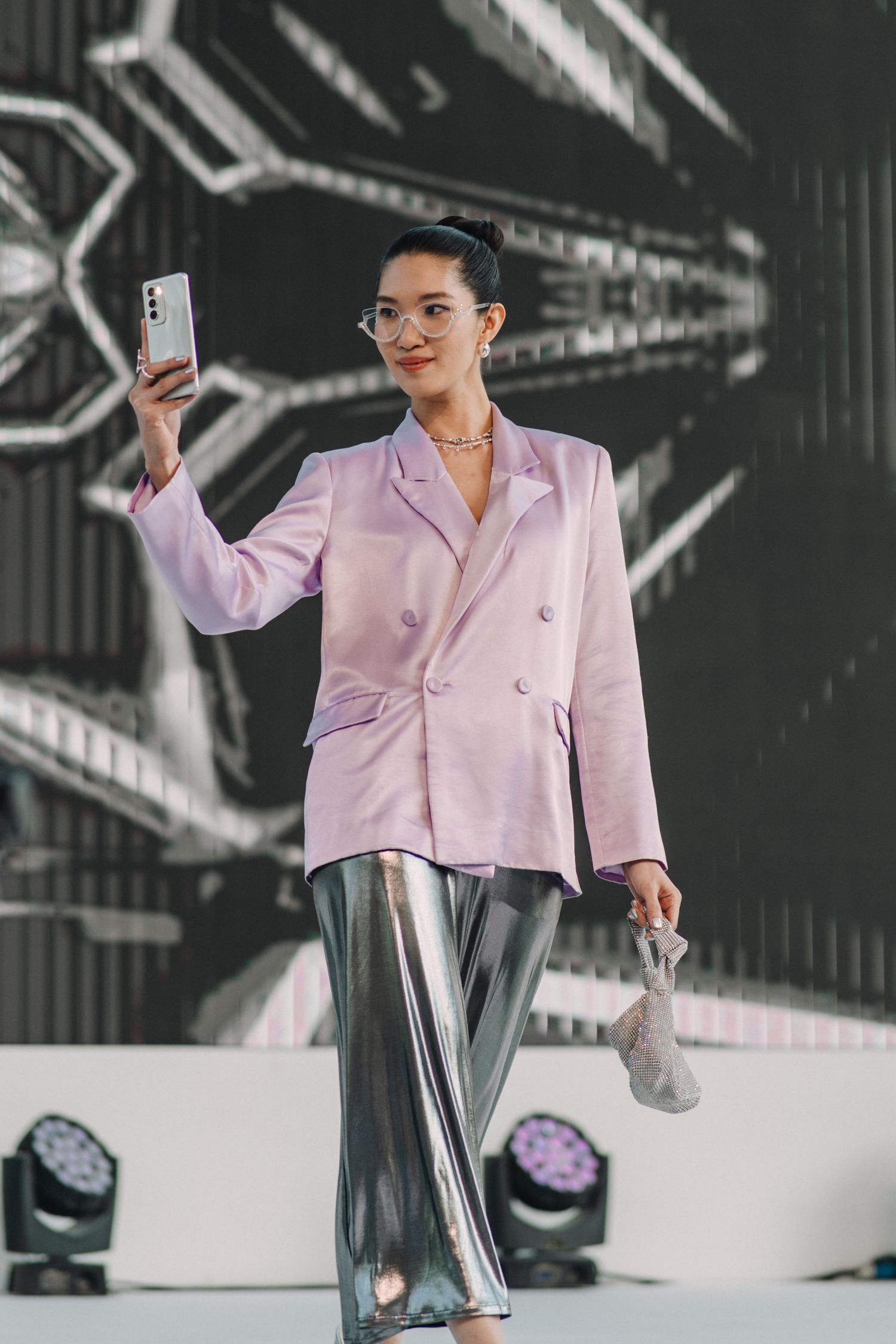 Fashion model in pink blazer and metallic pants holding phone on modern runway stage.