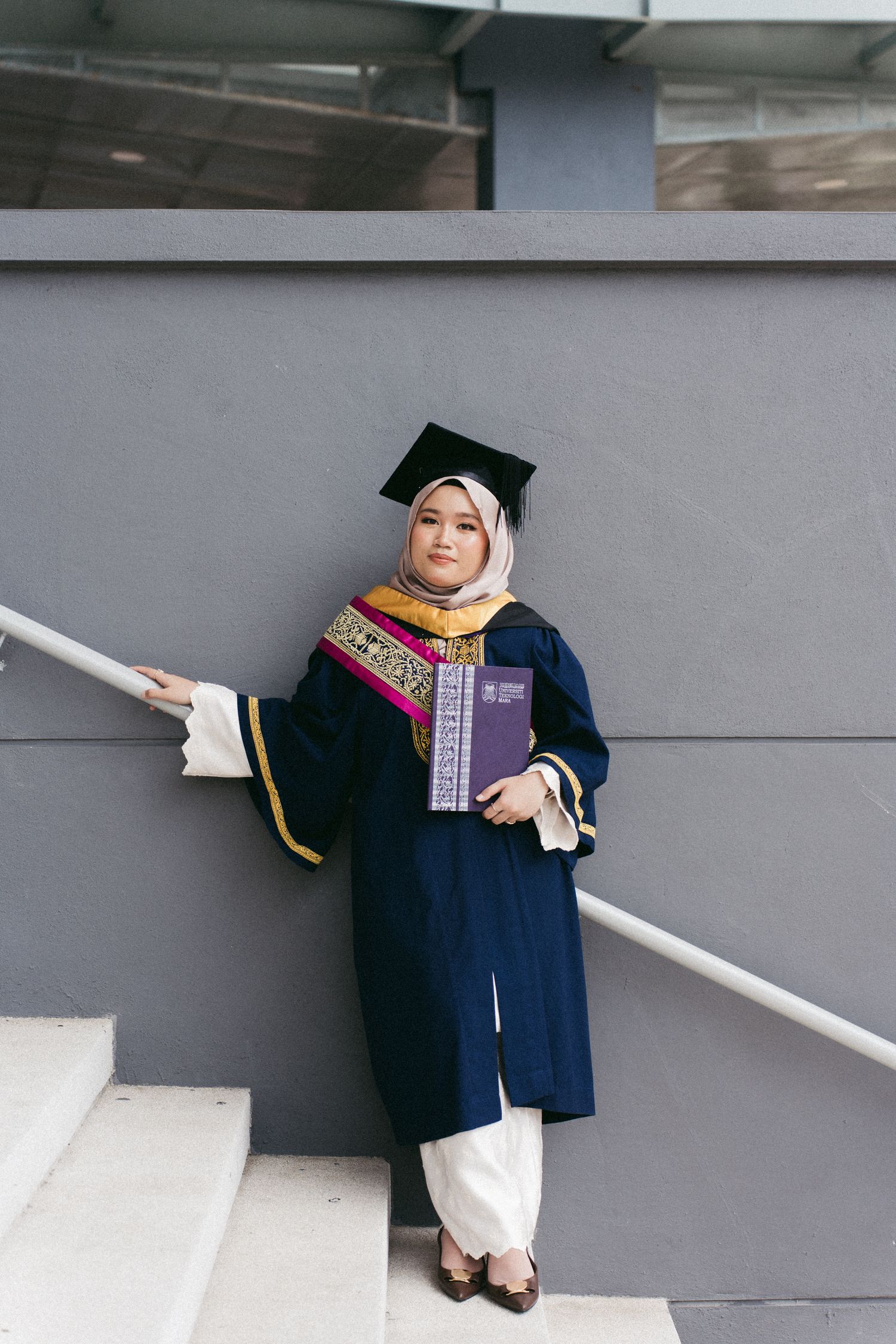 A graduate in traditional hijab and navy graduation gown poses on gray steps, wearing academic regalia and stole.