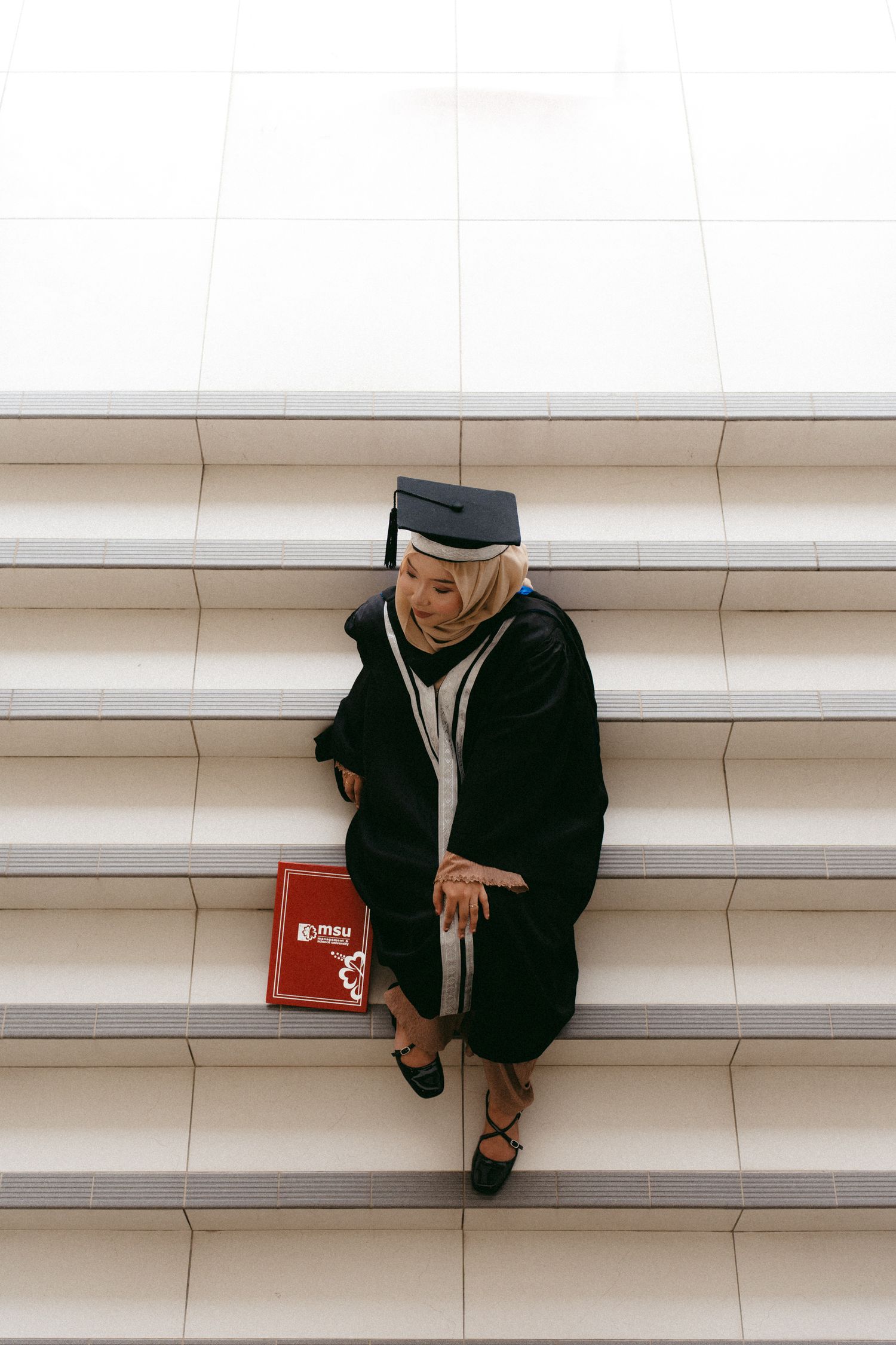 A graduate student sits alone on white stairs holding a red book while wearing academic regalia in black.