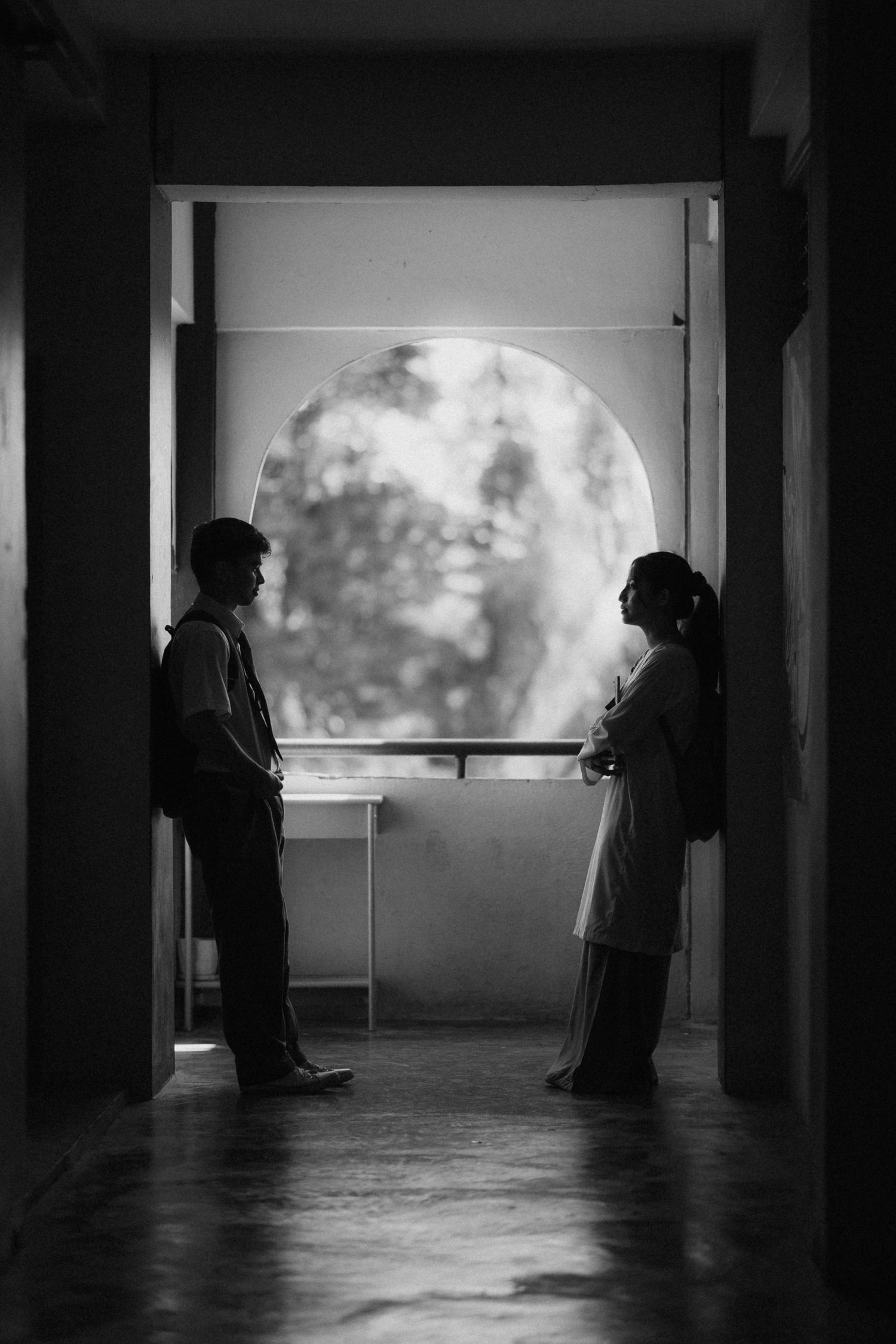 Two silhouetted figures stand in a dark hallway facing each other in front of a bright circular window.
