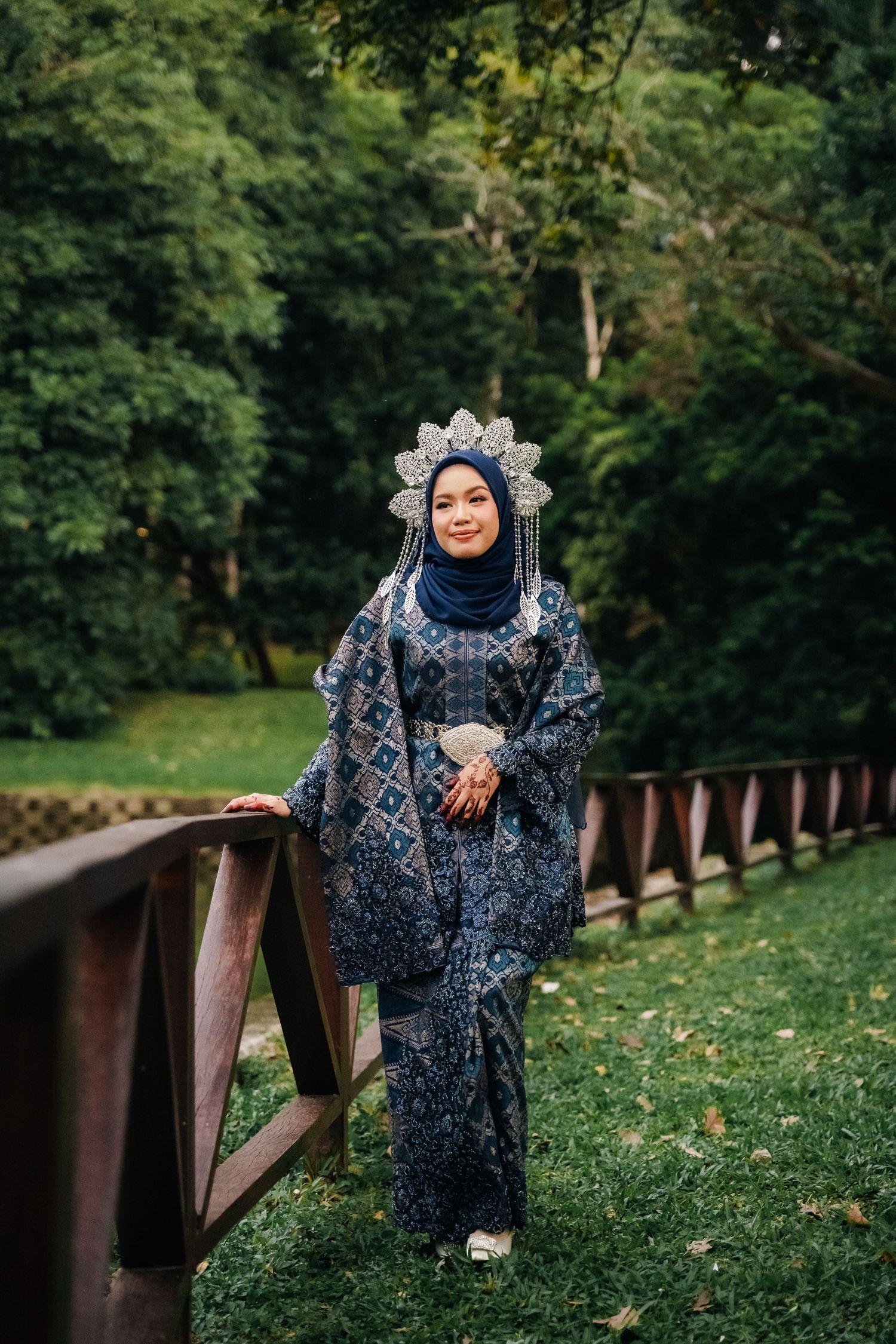 A person in a traditional blue patterned dress and headpiece poses on a wooden bridge surrounded by lush greenery.