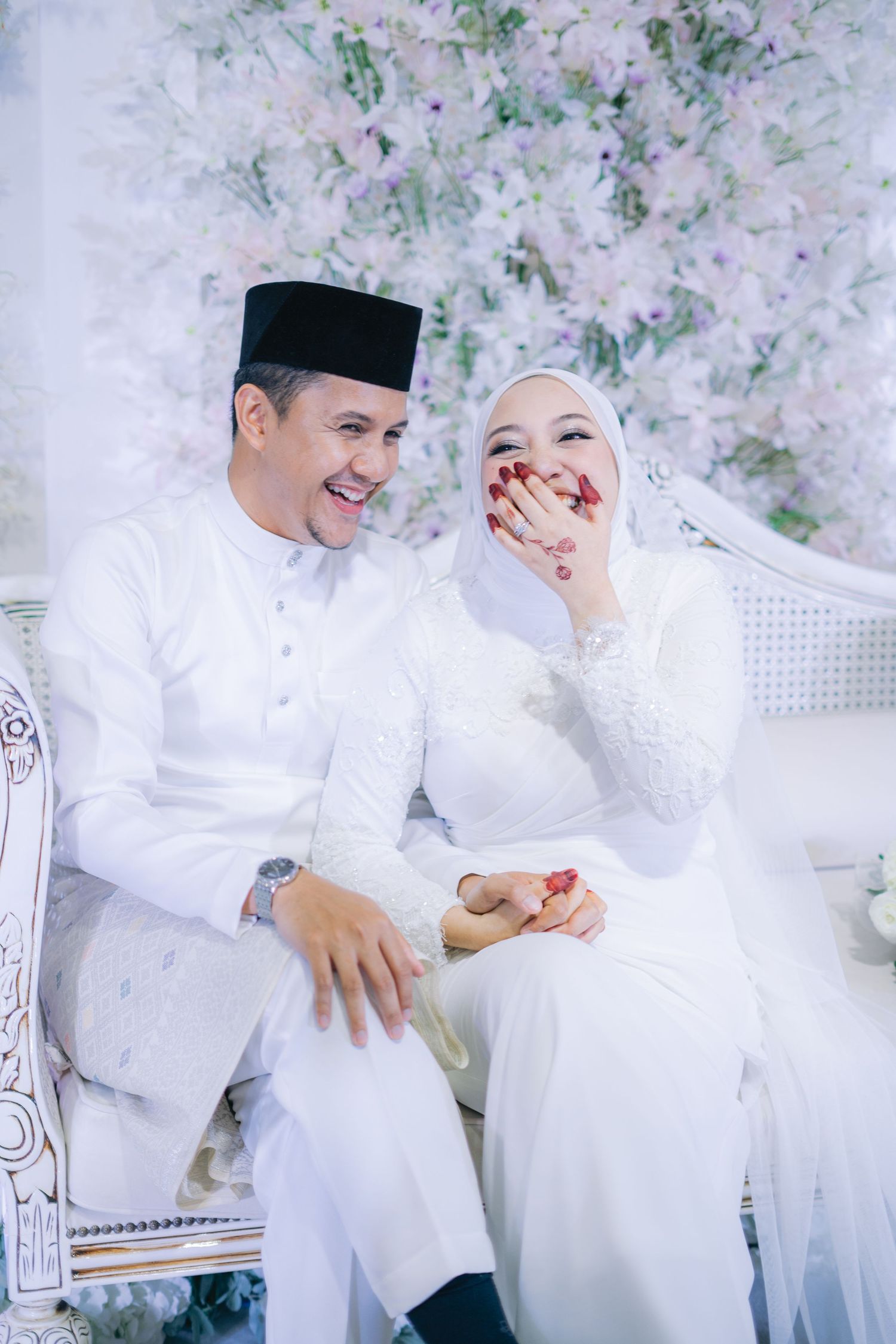 A couple in traditional white Malaysian wedding attire share a joyful moment on an ornate chair against a floral backdrop.
