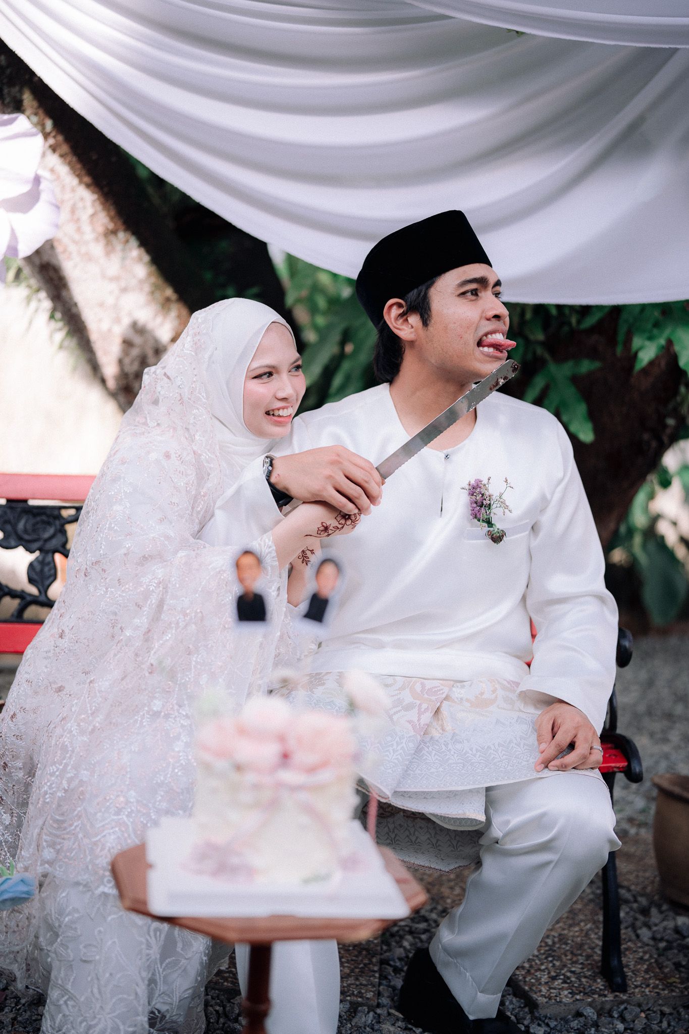 Muslim couple in white traditional attire cutting wedding cake outdoors, bride in hijab feeding groom.
