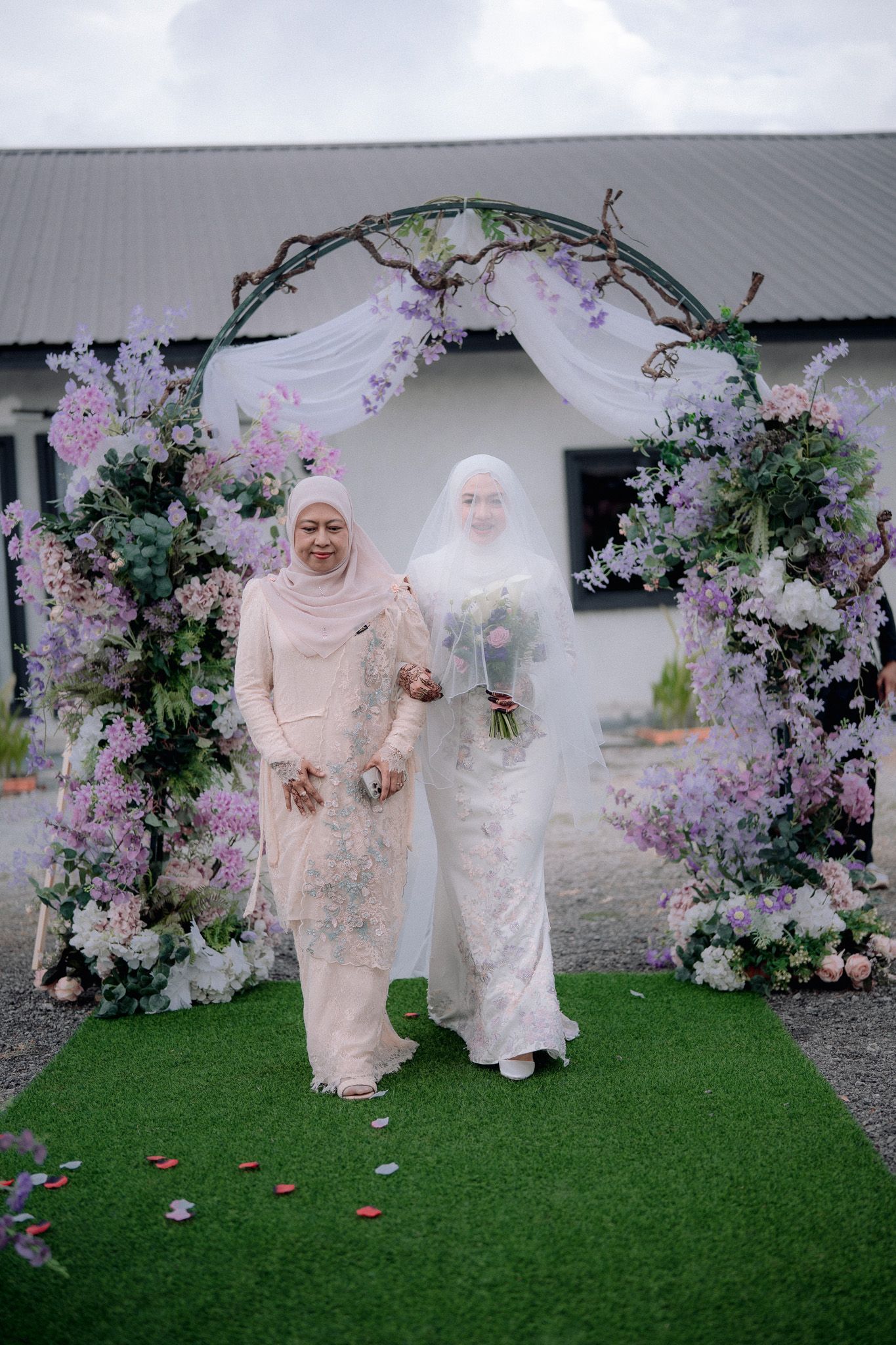 Muslim bride in white lace gown holding bouquet with elder woman under floral arch decorated with purple flowers.