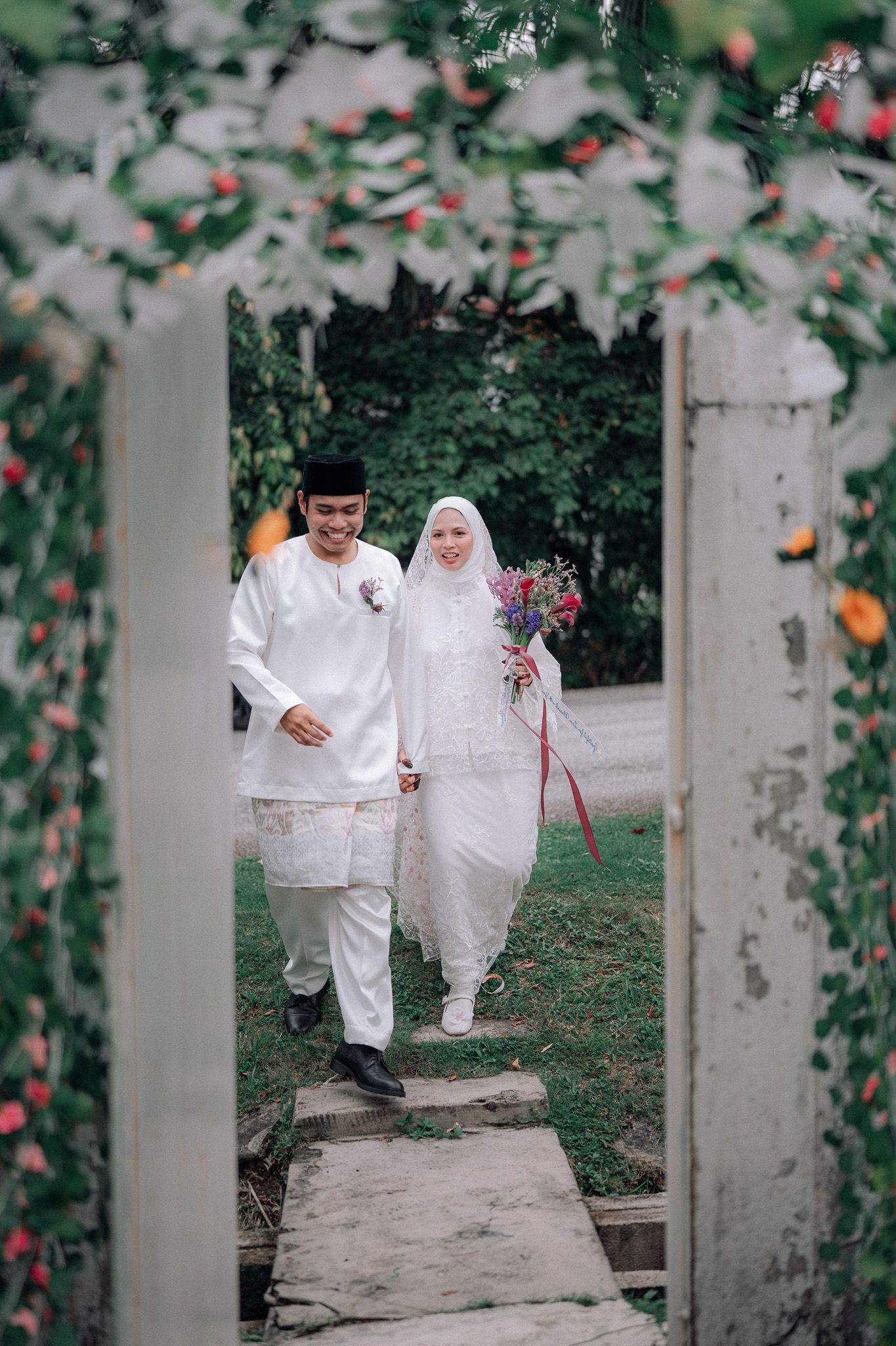 Muslim couple in white traditional attire walking through a floral arch on their wedding day, bride holding colorful bouquet.