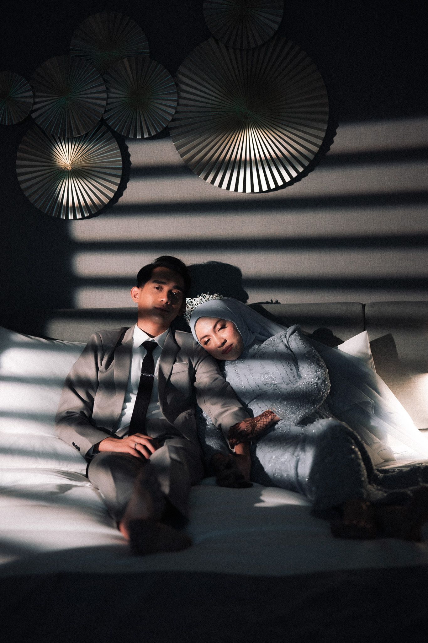 Muslim couple in wedding attire sitting on bed with dramatic shadow patterns and decorative fans on wall.