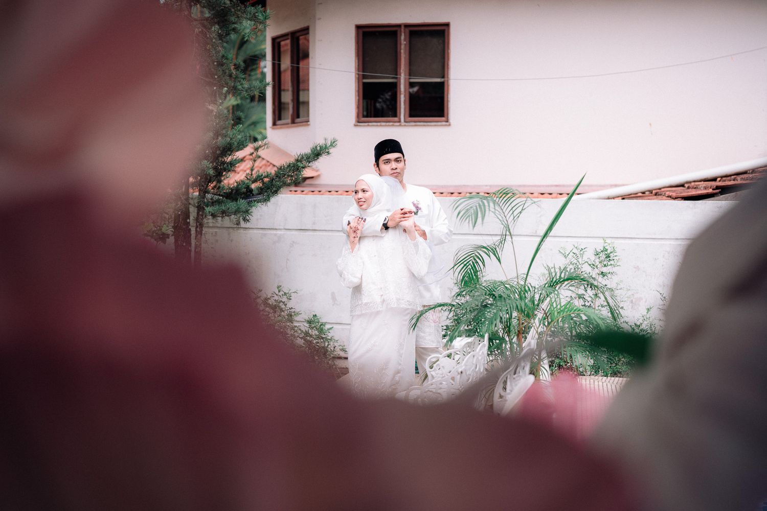 Muslim couple in white wedding attire standing together outdoors near a building with lush greenery.