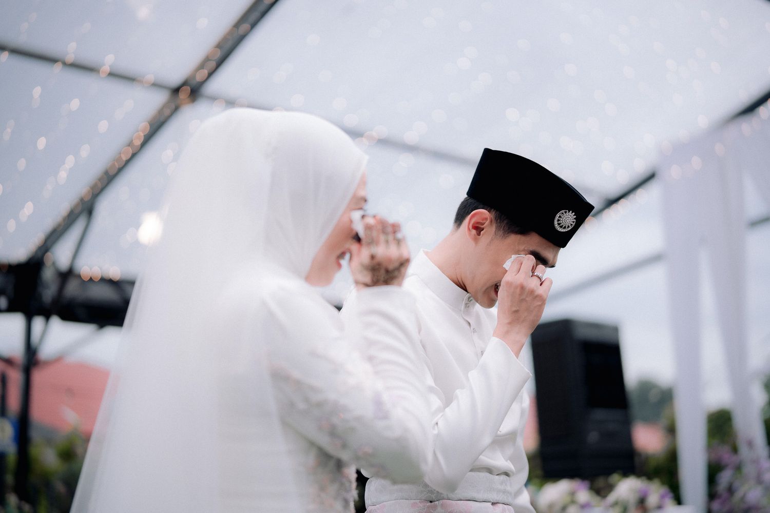 Muslim couple in white attire sharing an emotional wedding moment, bride in hijab and groom in songkok cap.