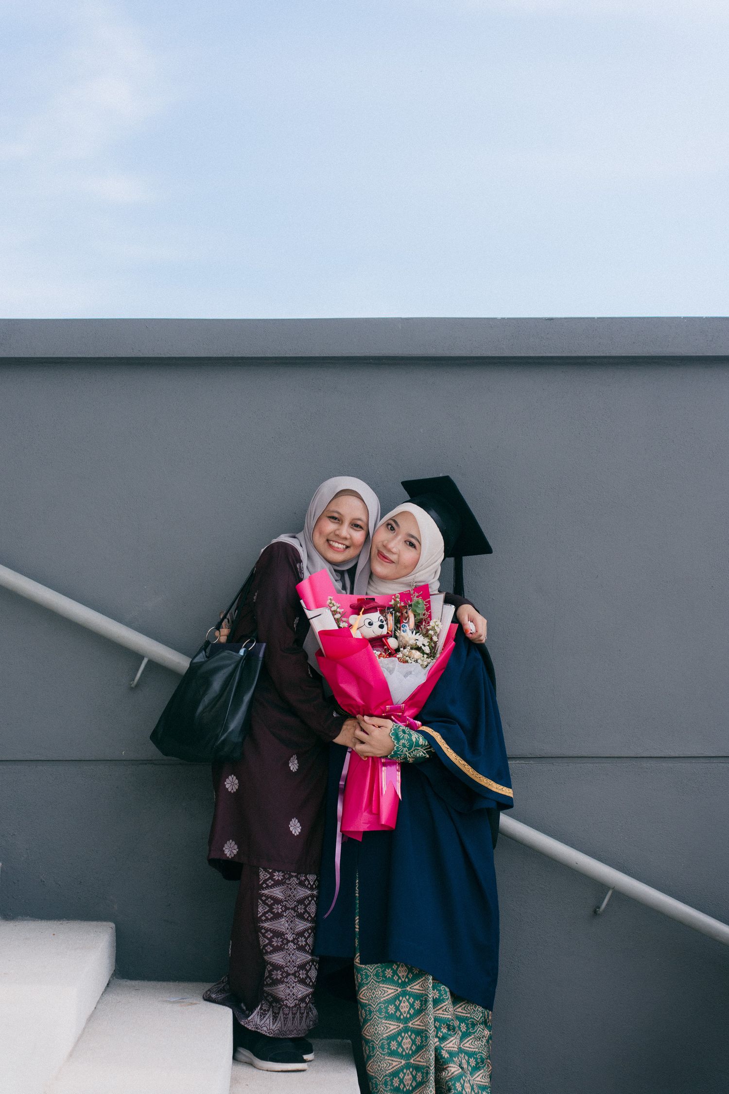 Two people in graduation attire lean against a gray wall while posing for photos.