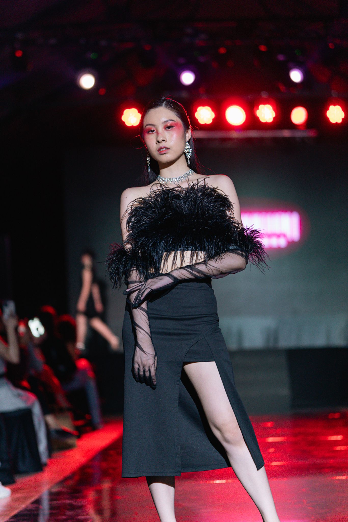 Model in black dress with feather detail poses dramatically on red-lit fashion runway.