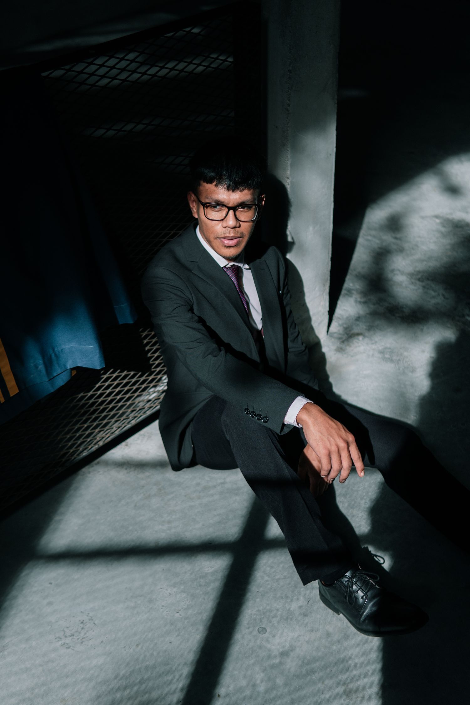 A person in formal business attire sits in dramatic lighting creating strong shadows.