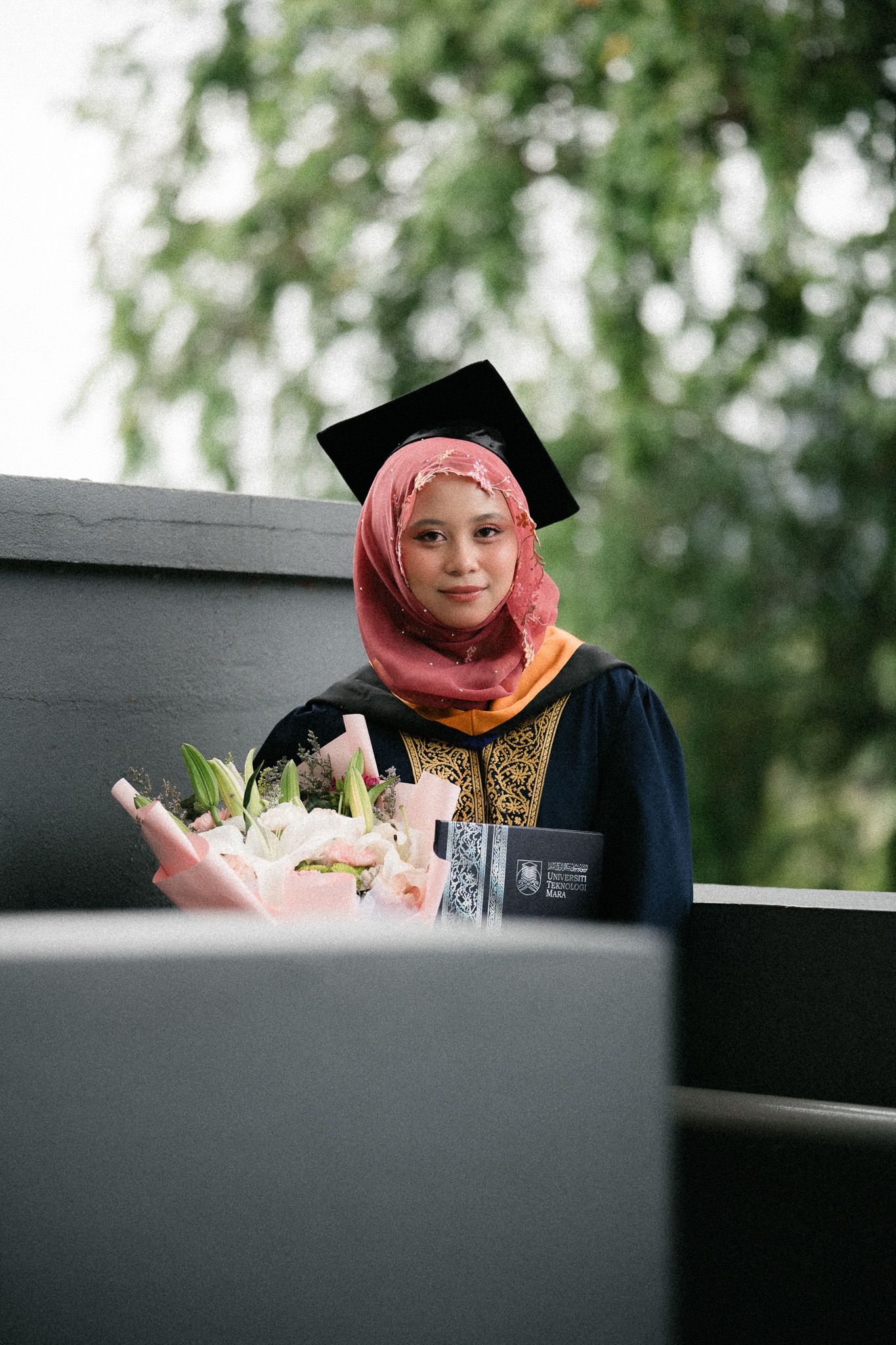 A young woman in graduation cap, gown, and pink hijab holds a bouquet of flowers while posing outdoors.